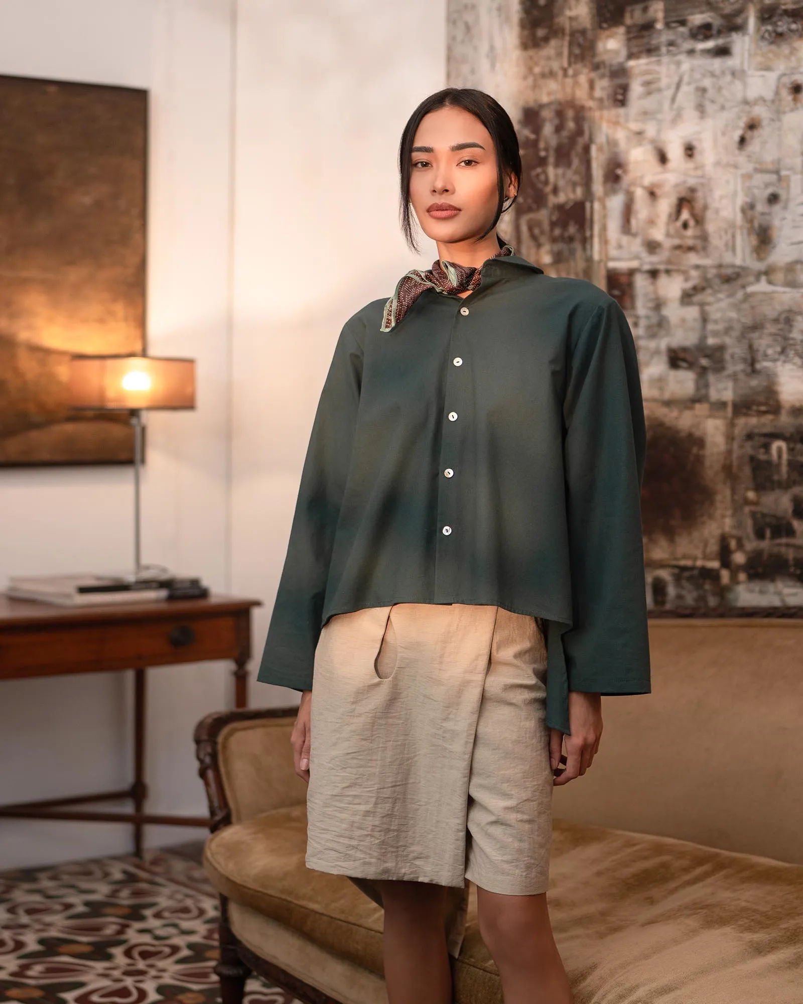 Person standing in a living room wearing a dark green button-down shirt, beige shorts, and a patterned scarf at the neck.