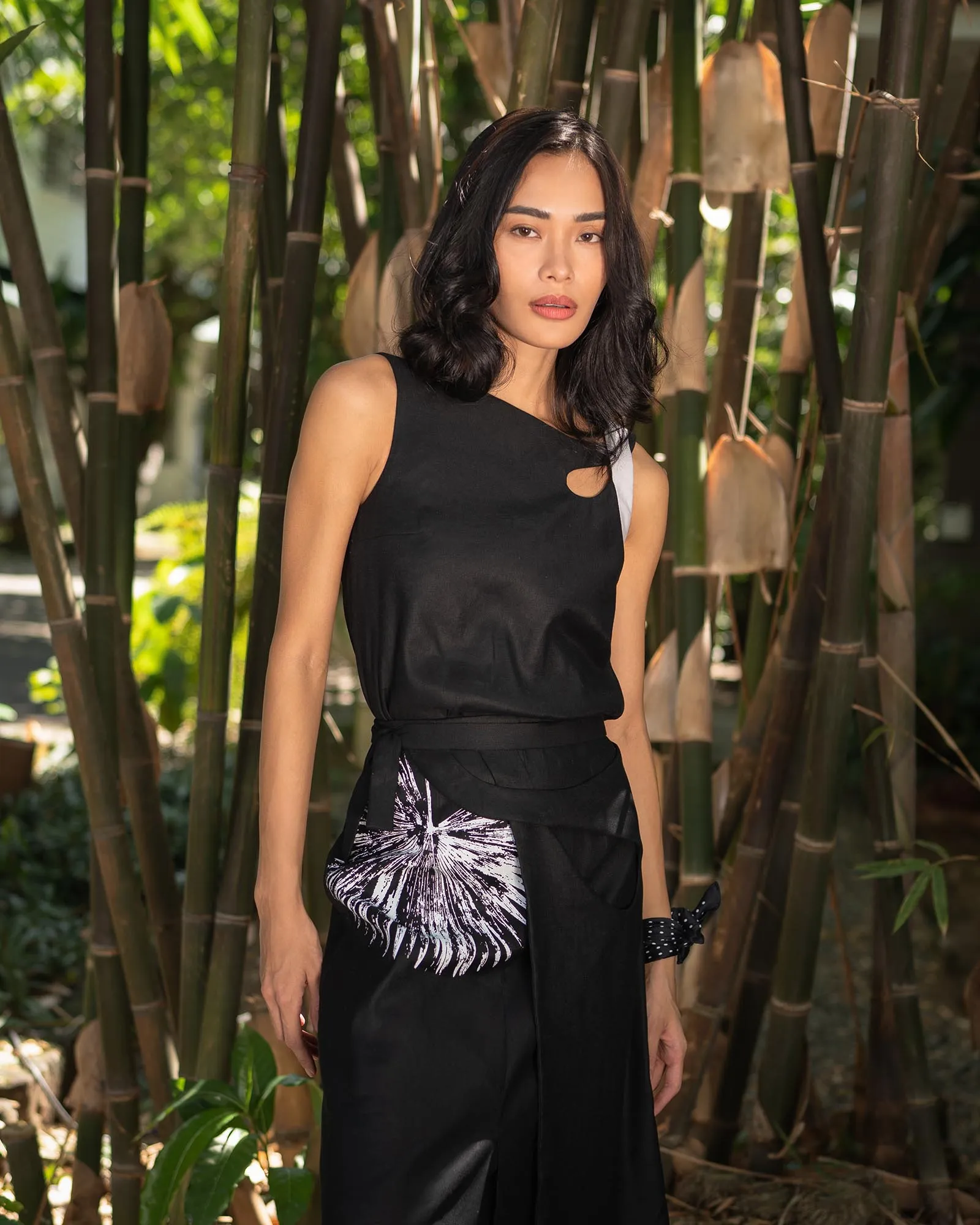 Woman with shoulder-length black hair wearing a sleeveless black dress stands among tall bamboo stalks in a sunny outdoor setting. This portrait highlights the model amid lush bamboo.