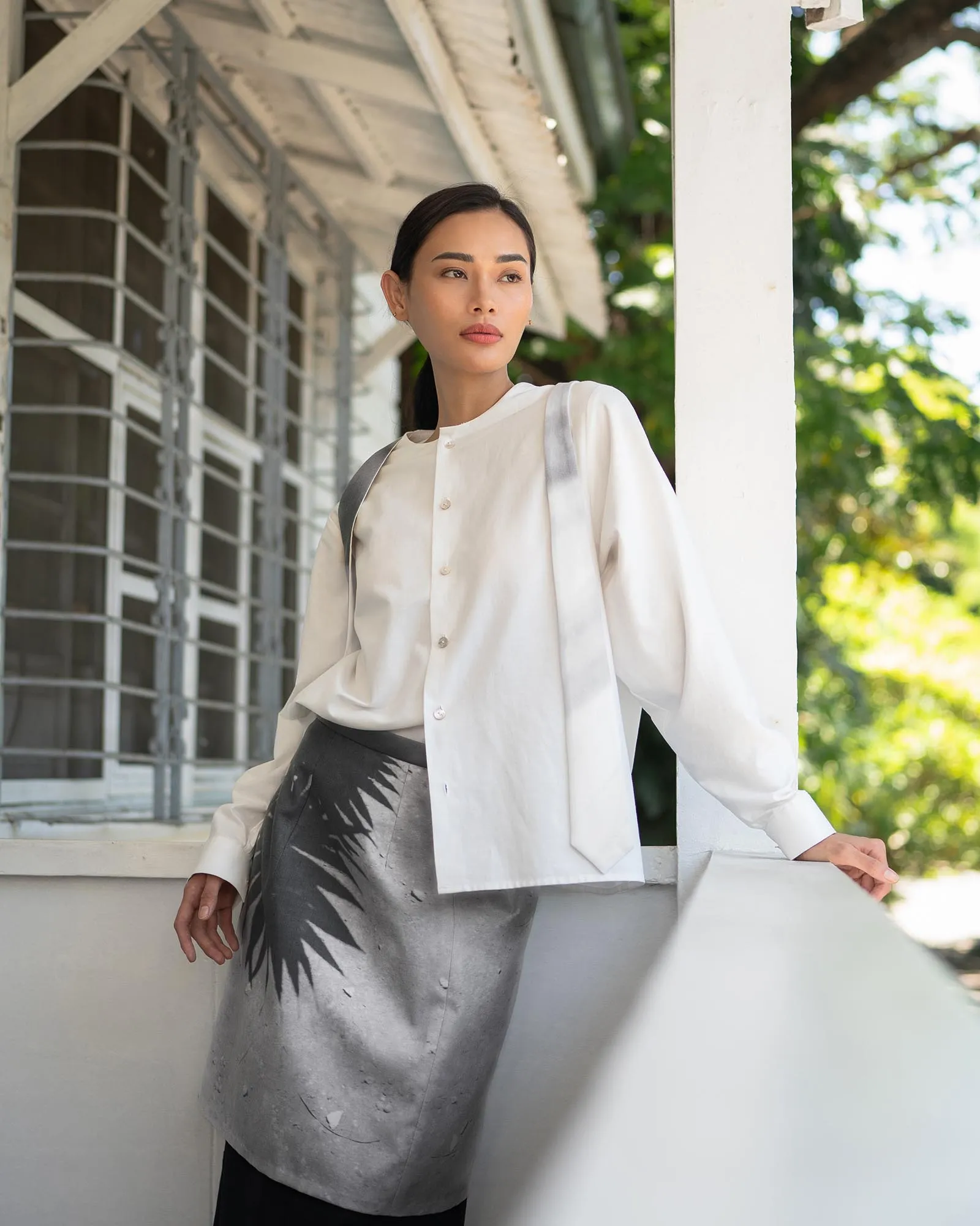 Woman leaning on a white railing outdoors, wearing a light beige button-up shirt over a patterned gray and black skirt with a bag strap over her shoulder.