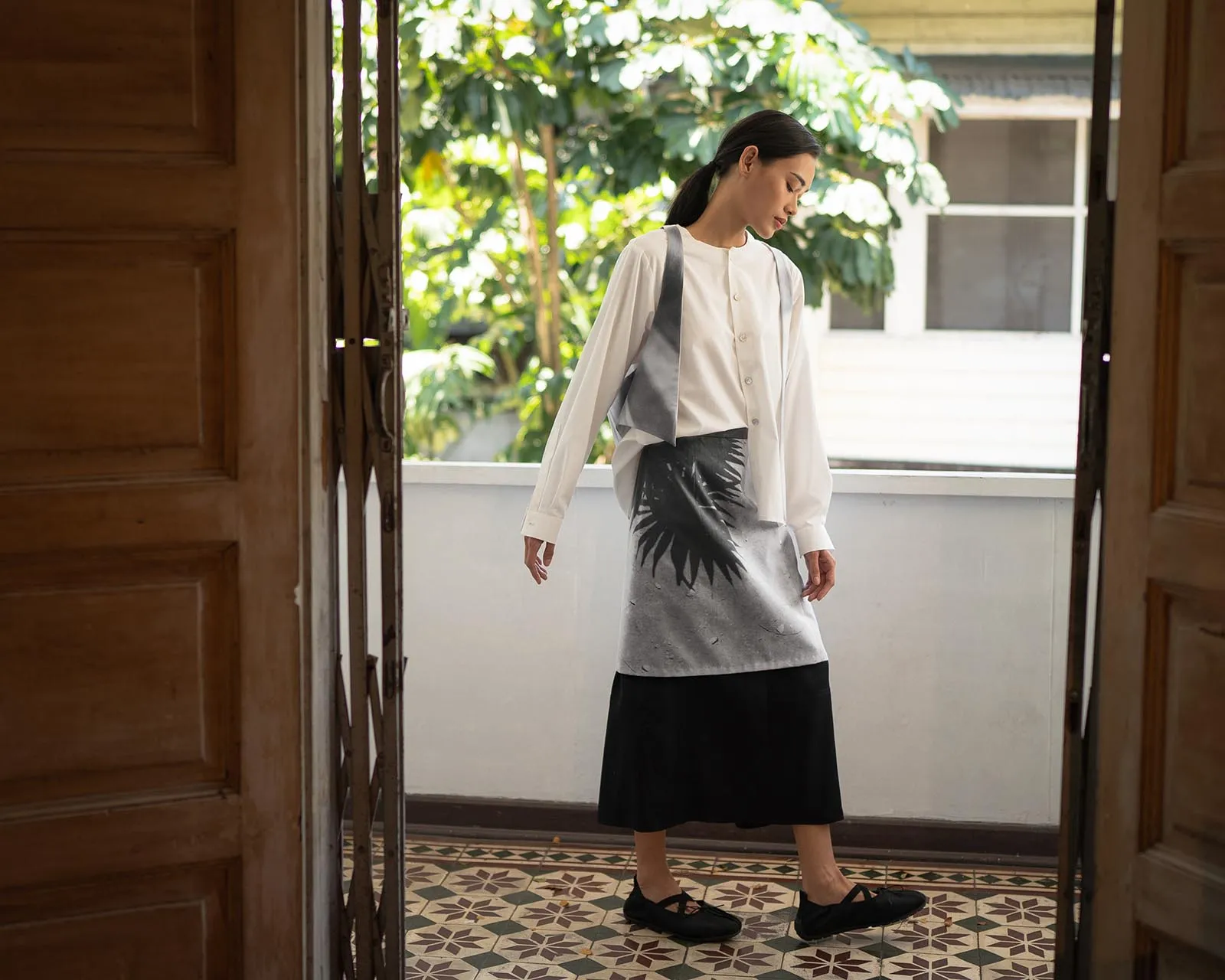 Woman standing in an open doorway, wearing a white blouse and a gray palm‑print dress with a black skirt, looking down; greenery outside the doorway.