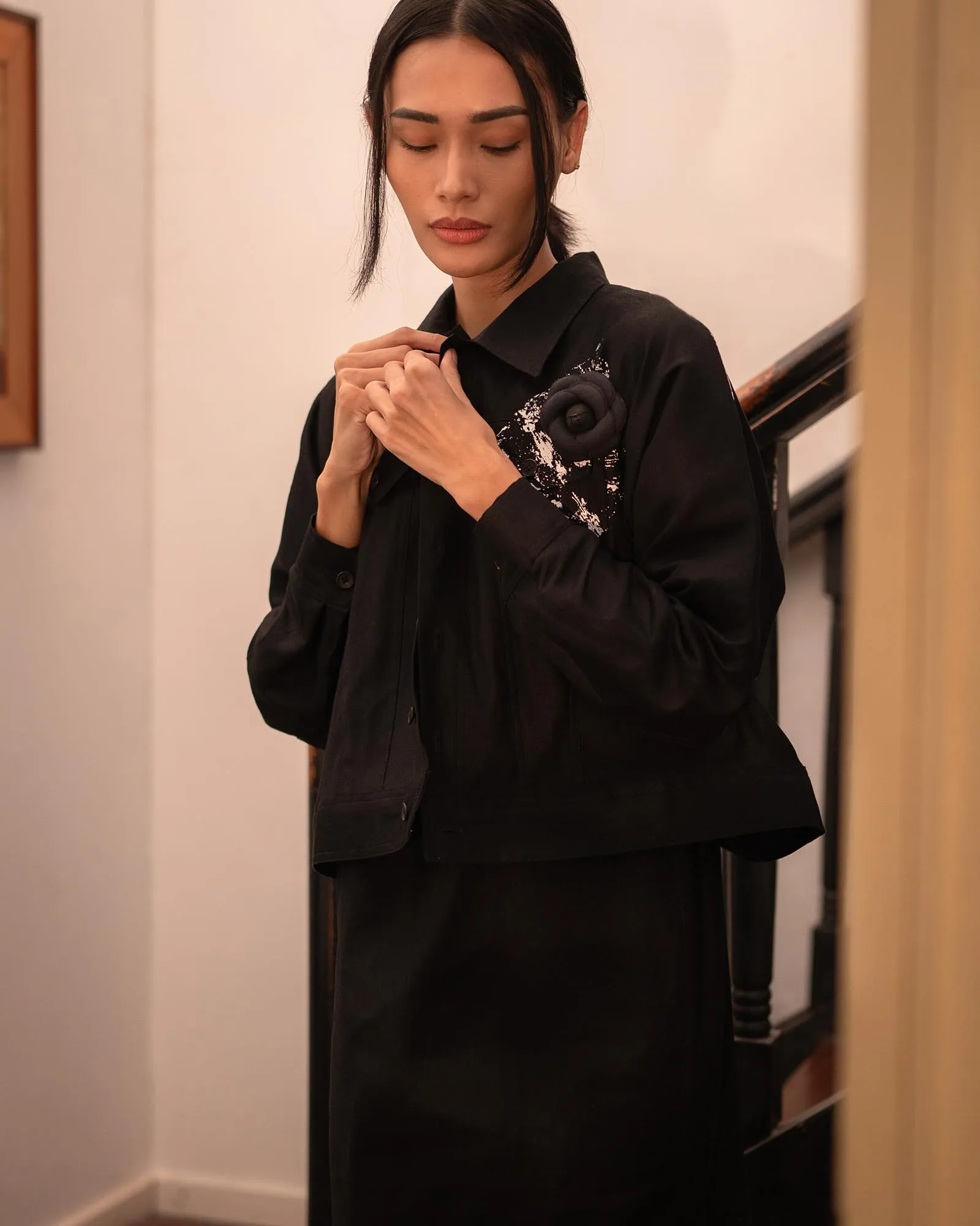 Woman in a black blazer adjusts her collar, with a black fabric flower pin on the chest, standing in a hallway beside a staircase.