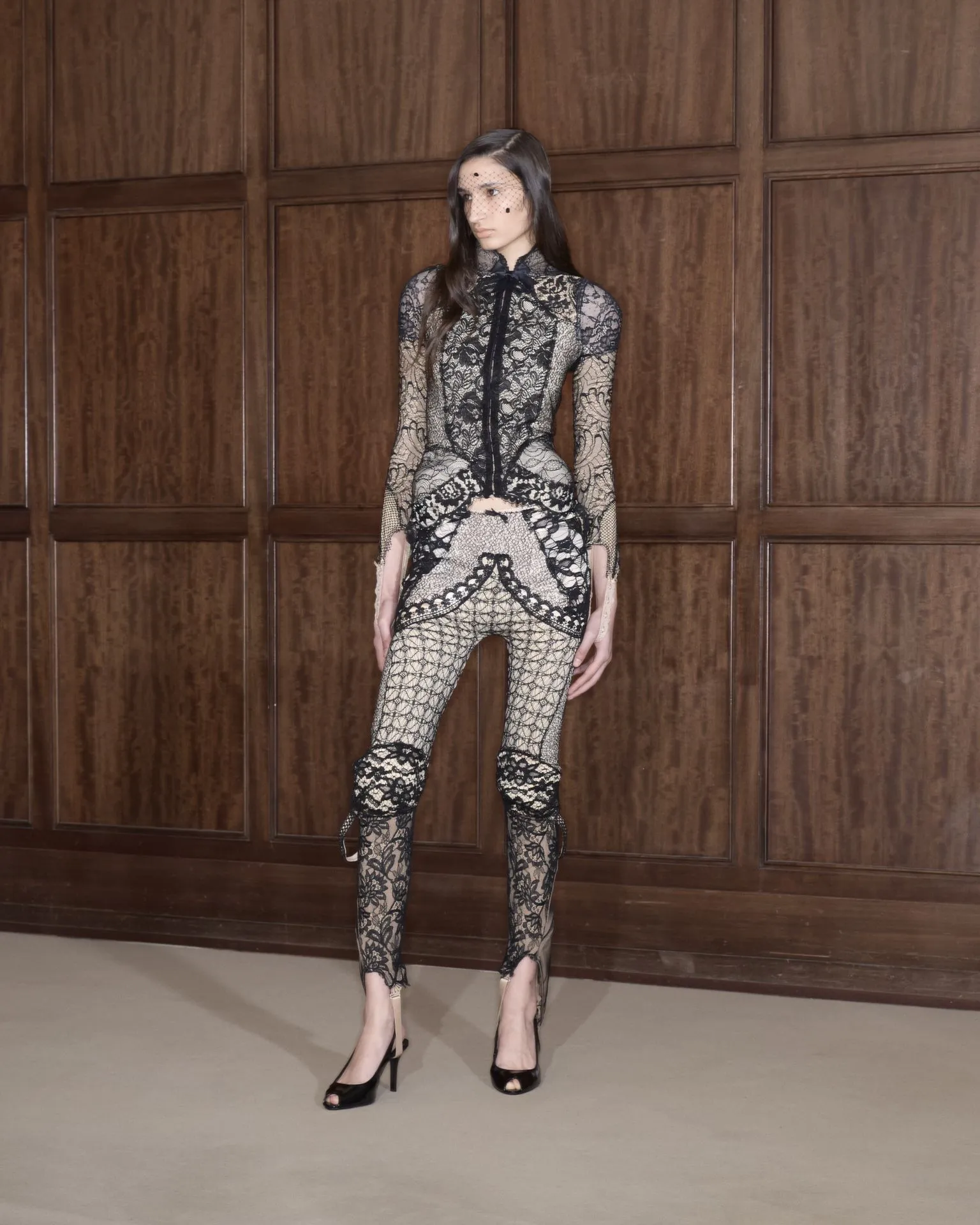 Full-body shot of a model in a black lace bodysuit with sheer panels, standing against dark wood paneling in high heels.