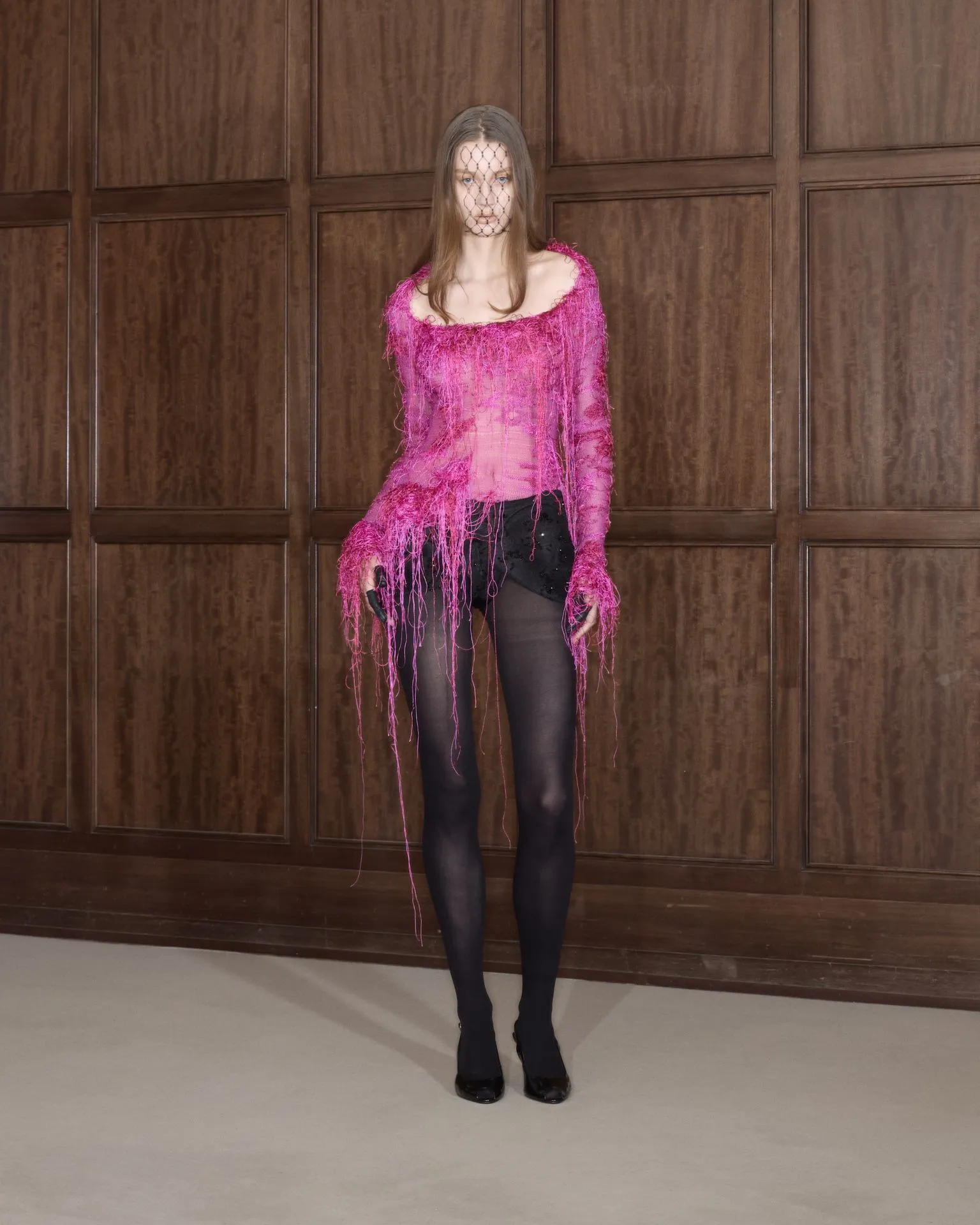 Woman wearing a bright pink, fringe-covered top with a net veil over her face, standing against dark wood paneled walls indoors.