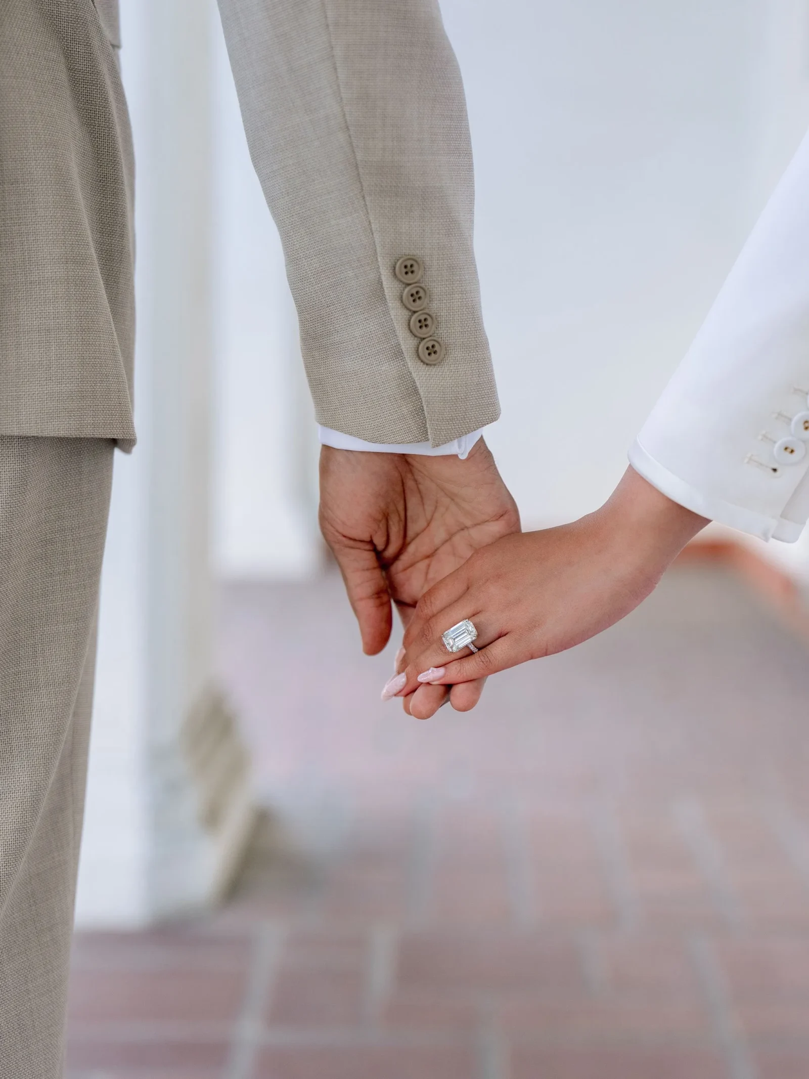 Couple holding hands; man in a beige suit sleeve and a woman in a white sleeve, ring visible on her finger.
