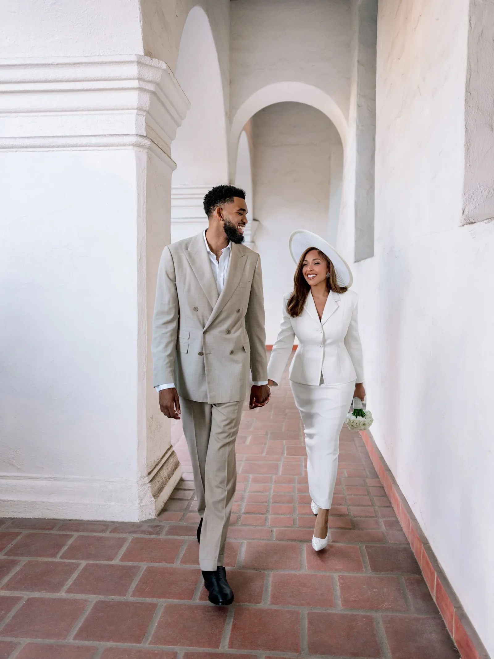 Man in a beige suit and a woman in a white suit walk hand in hand through a light, arched walkway; she carries a small bouquet of white flowers.