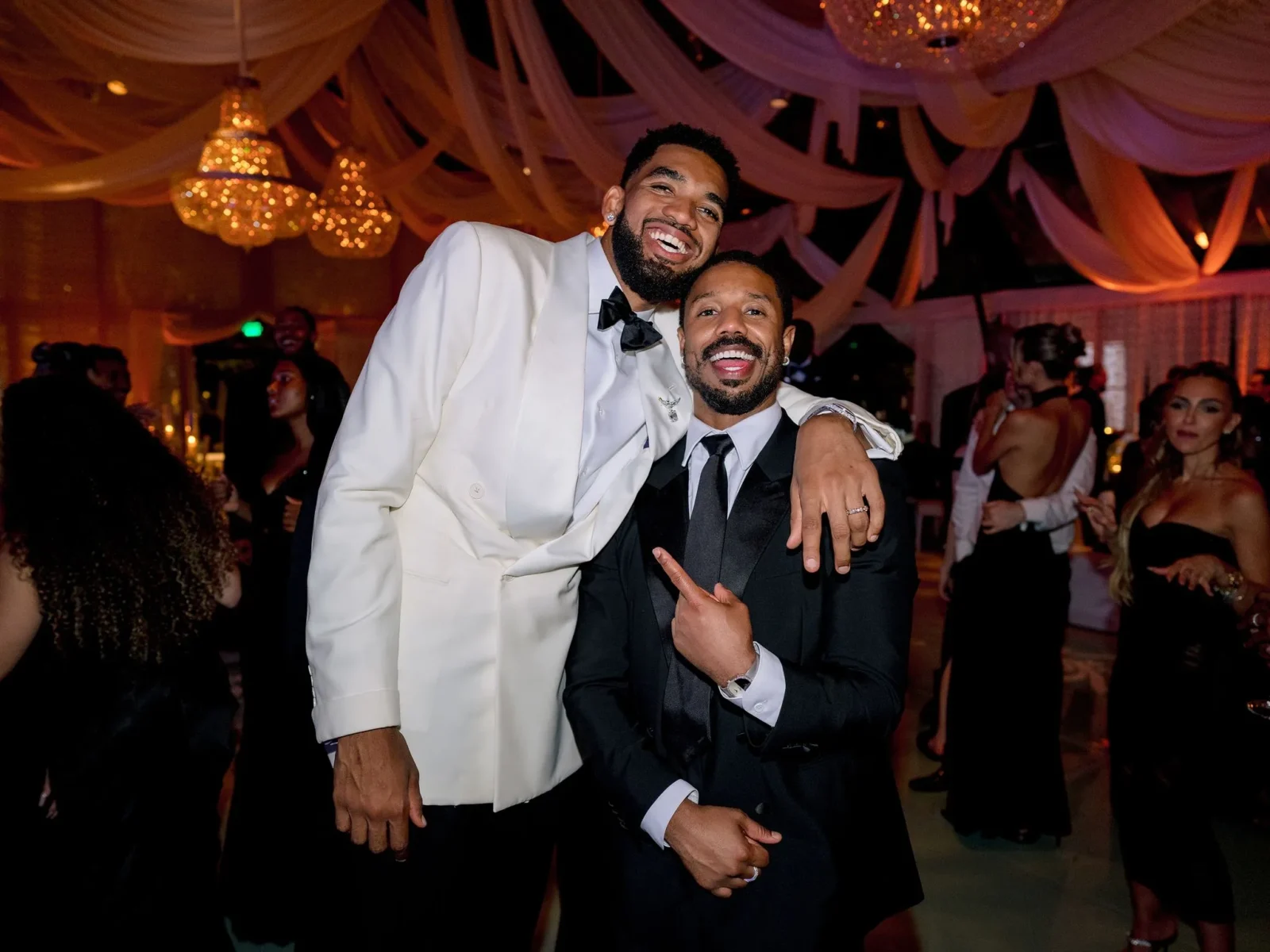 Two men in tuxedos posing and smiling at a festive, chandelier-lit event with draped white fabric overhead.