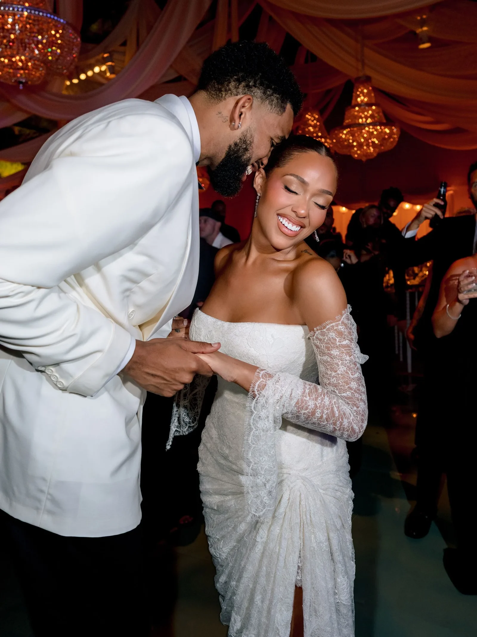 Bride in an off‑the‑shoulder lace wedding dress smiling as she dances with a man in a white suit at a wedding reception under chandeliers, holding hands.