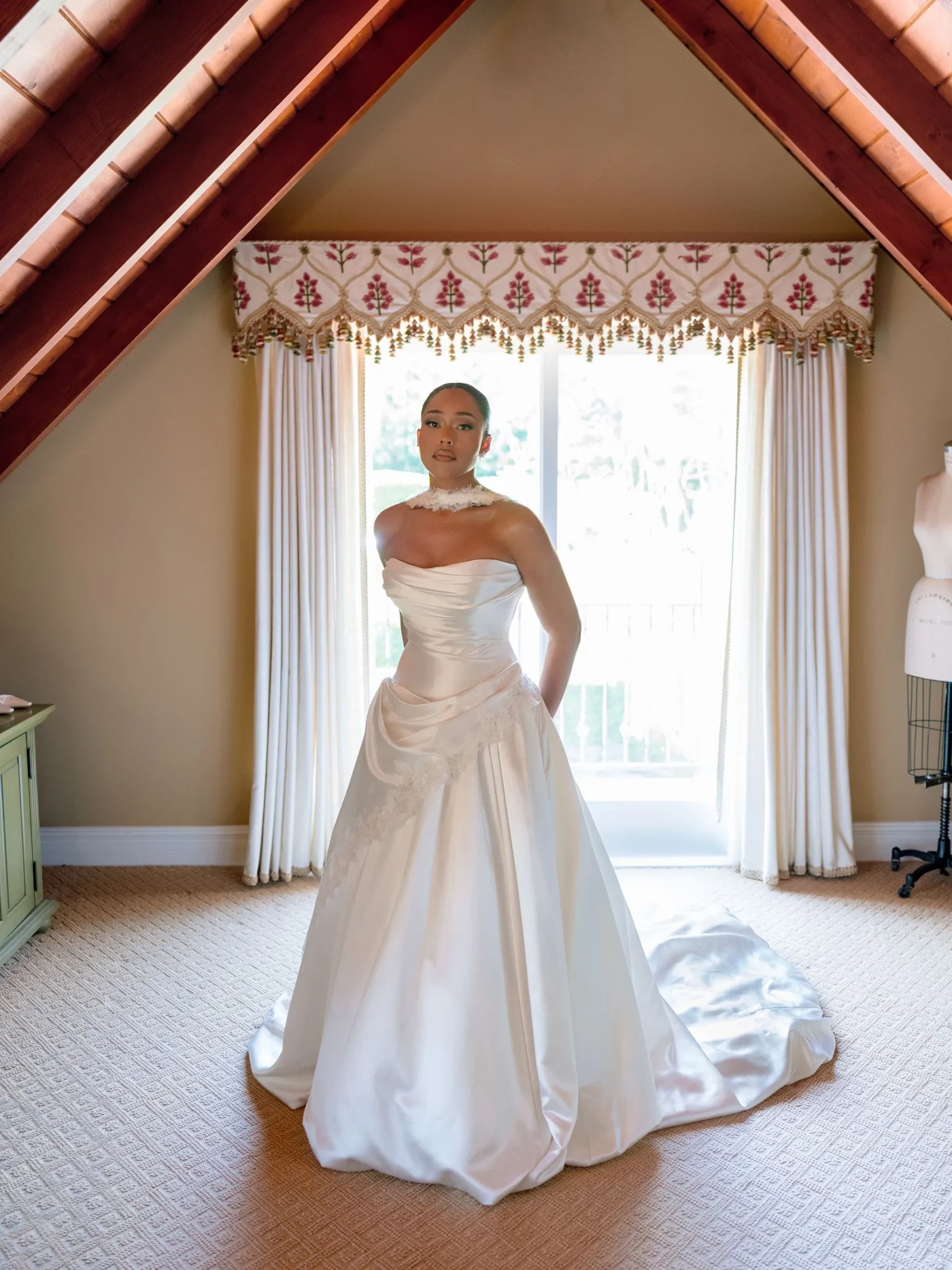 Bride in a white strapless wedding gown with lace accents stands in a sunlit room framed by slanted attic beams and a decorative valance above a window.