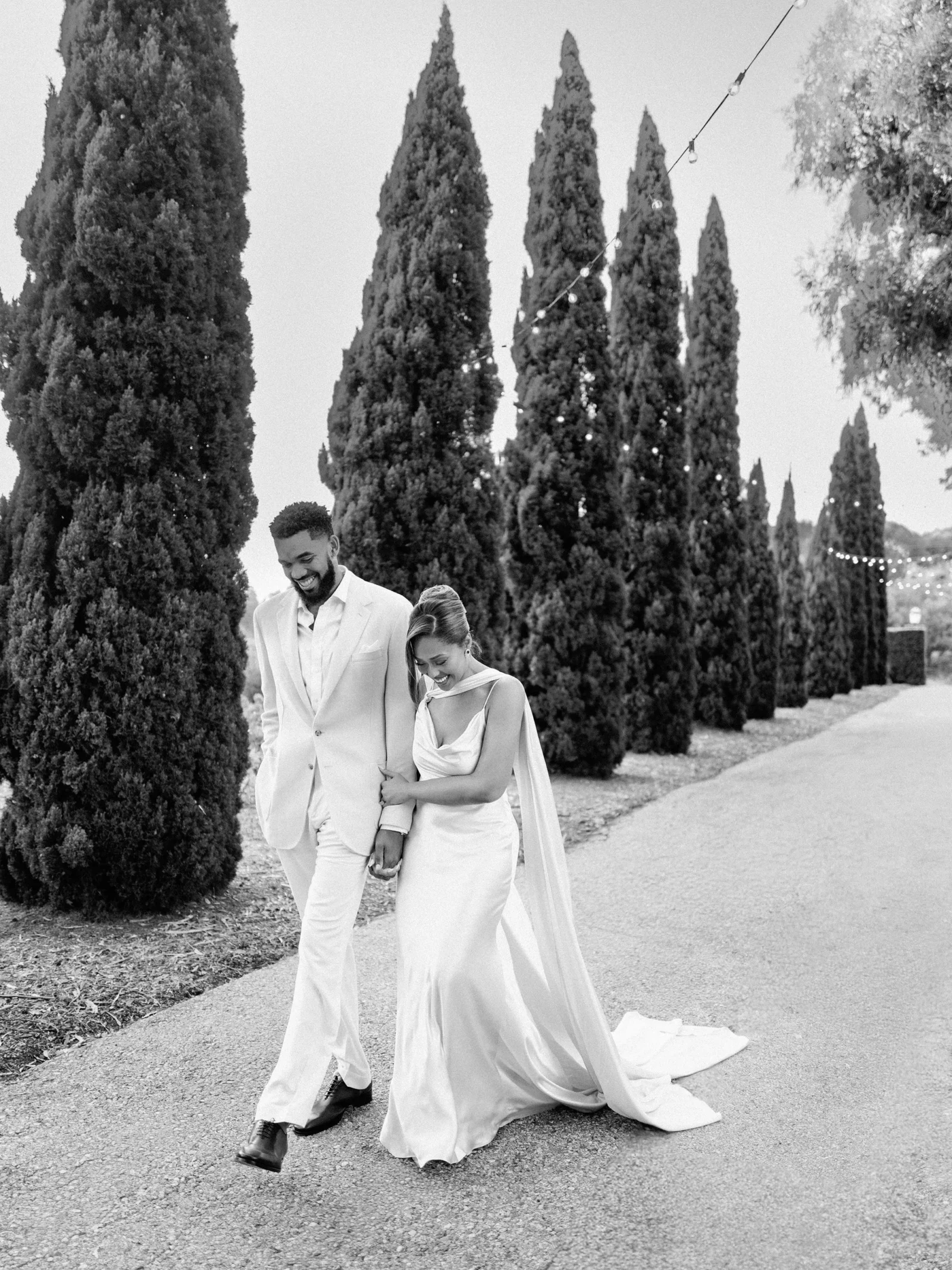 Bride and groom in wedding attire walk hand in hand down a gravel path lined with tall evergreen trees.