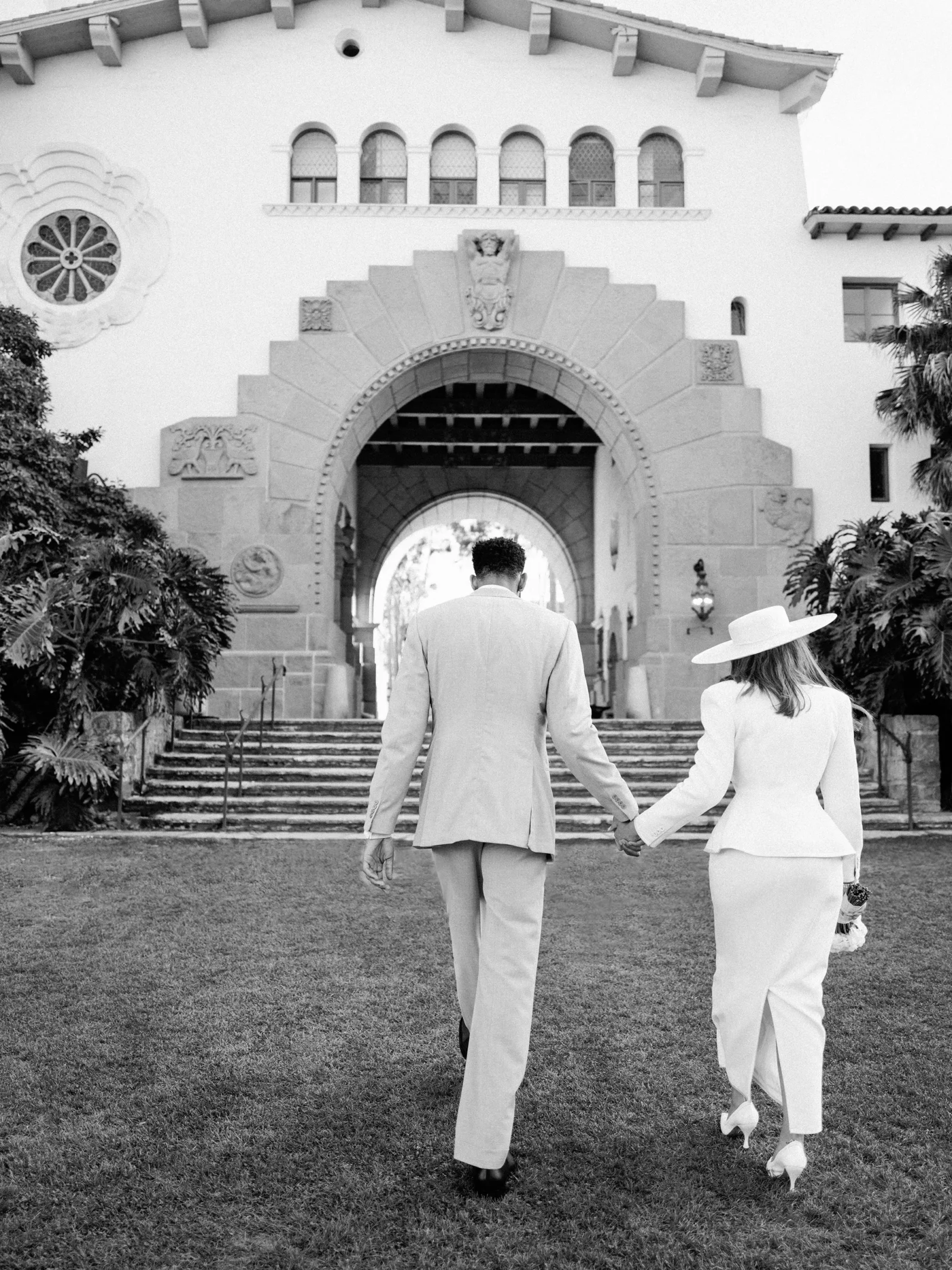 Couple in light formal attire walk hand in hand toward a large arched entrance of a historic-looking building in a black-and-white scene.