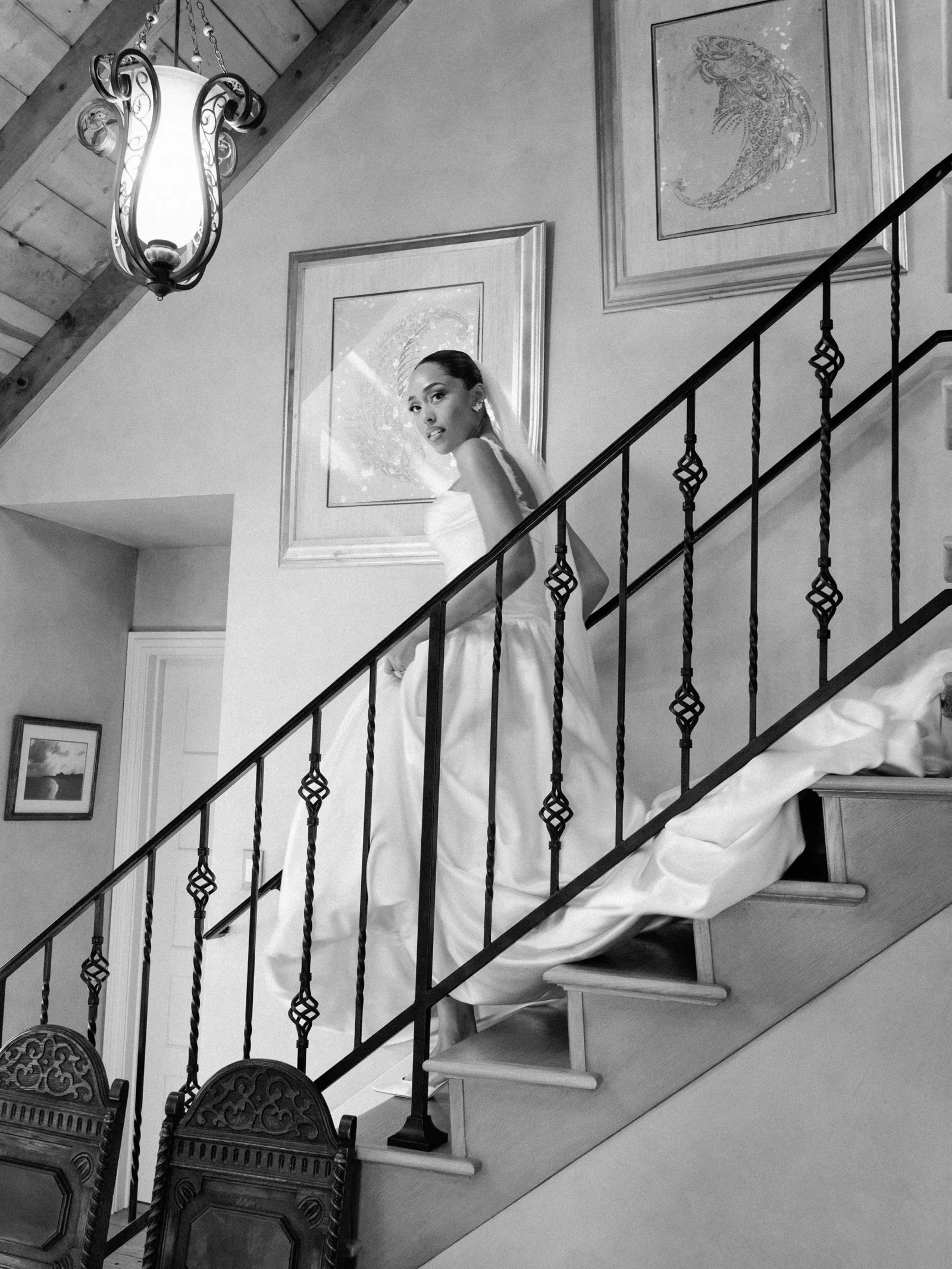 Bride in a wedding gown standing on a curved staircase, looking back over her shoulder toward the camera, ornate iron railing and framed artwork on the wall nearby.