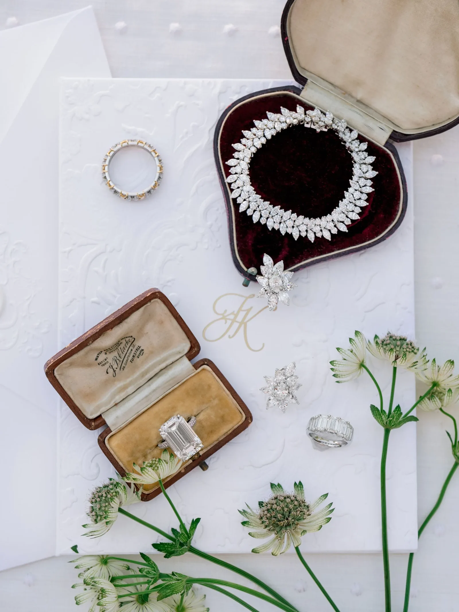 Diamond necklace with sparkling stones displayed in a burgundy velvet jewelry box, beside a ring in an open case and white flowers.