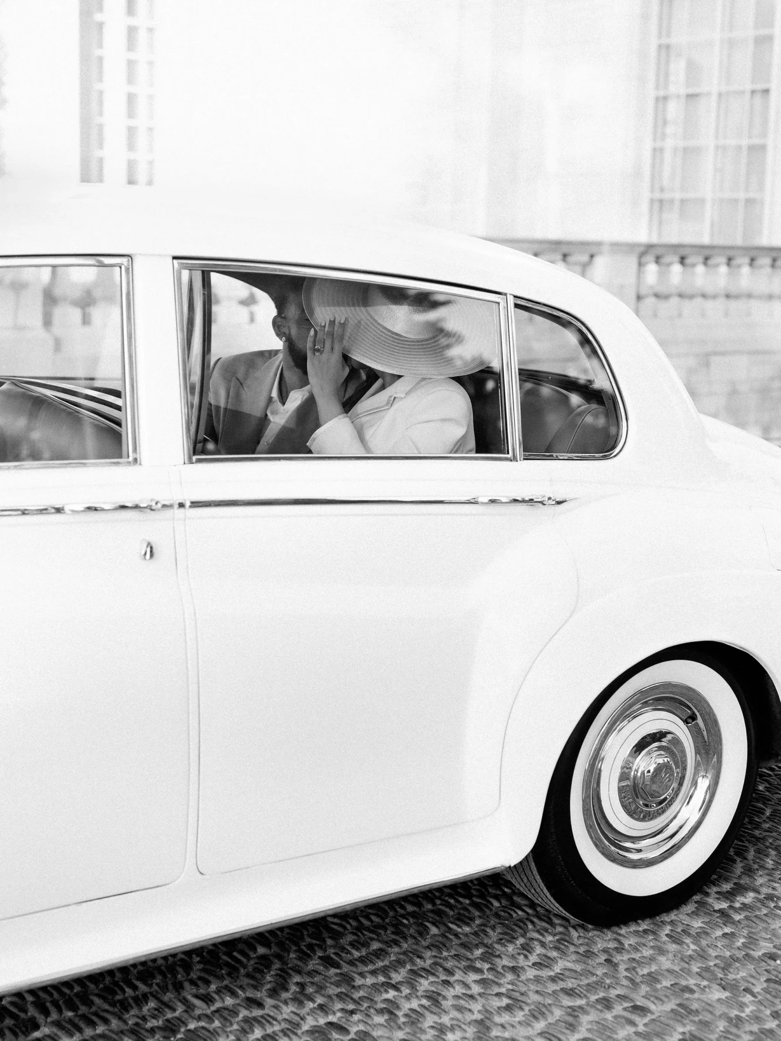Man and woman seated in a vintage white car, the woman wearing a wide-brimmed hat and touching her face through the window.