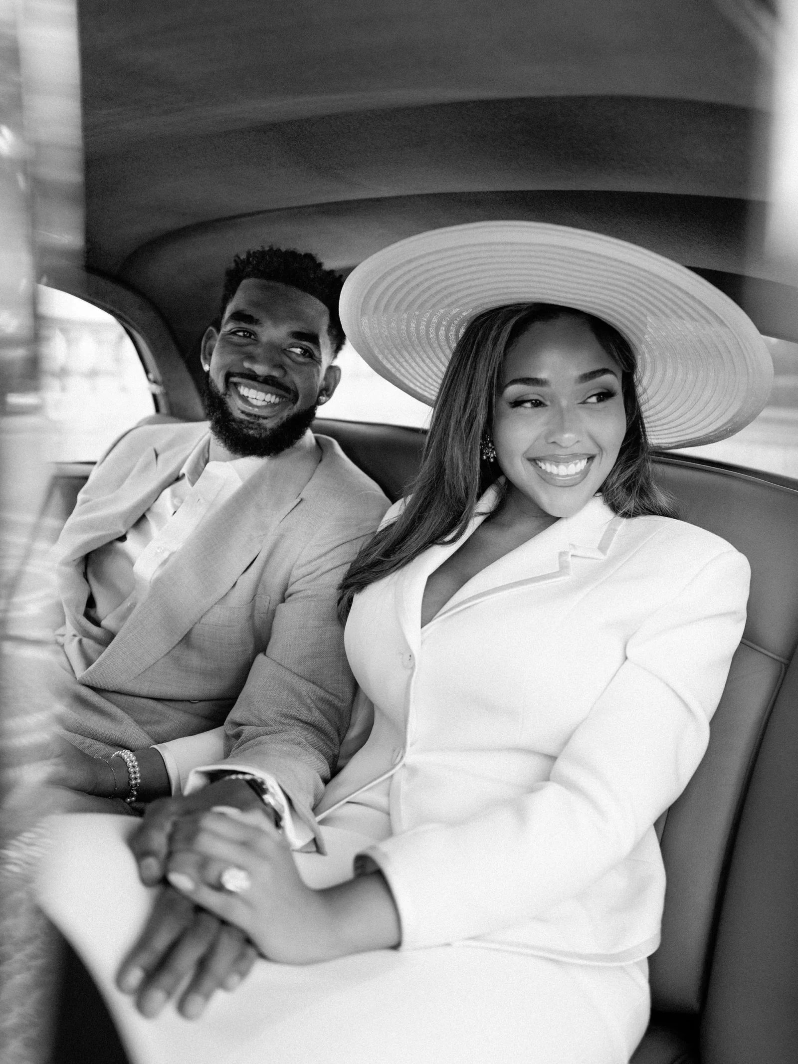 Smiling couple seated in a car backseat, the woman wearing a wide-brim hat and a light blazer beside a bearded man in a suit.
