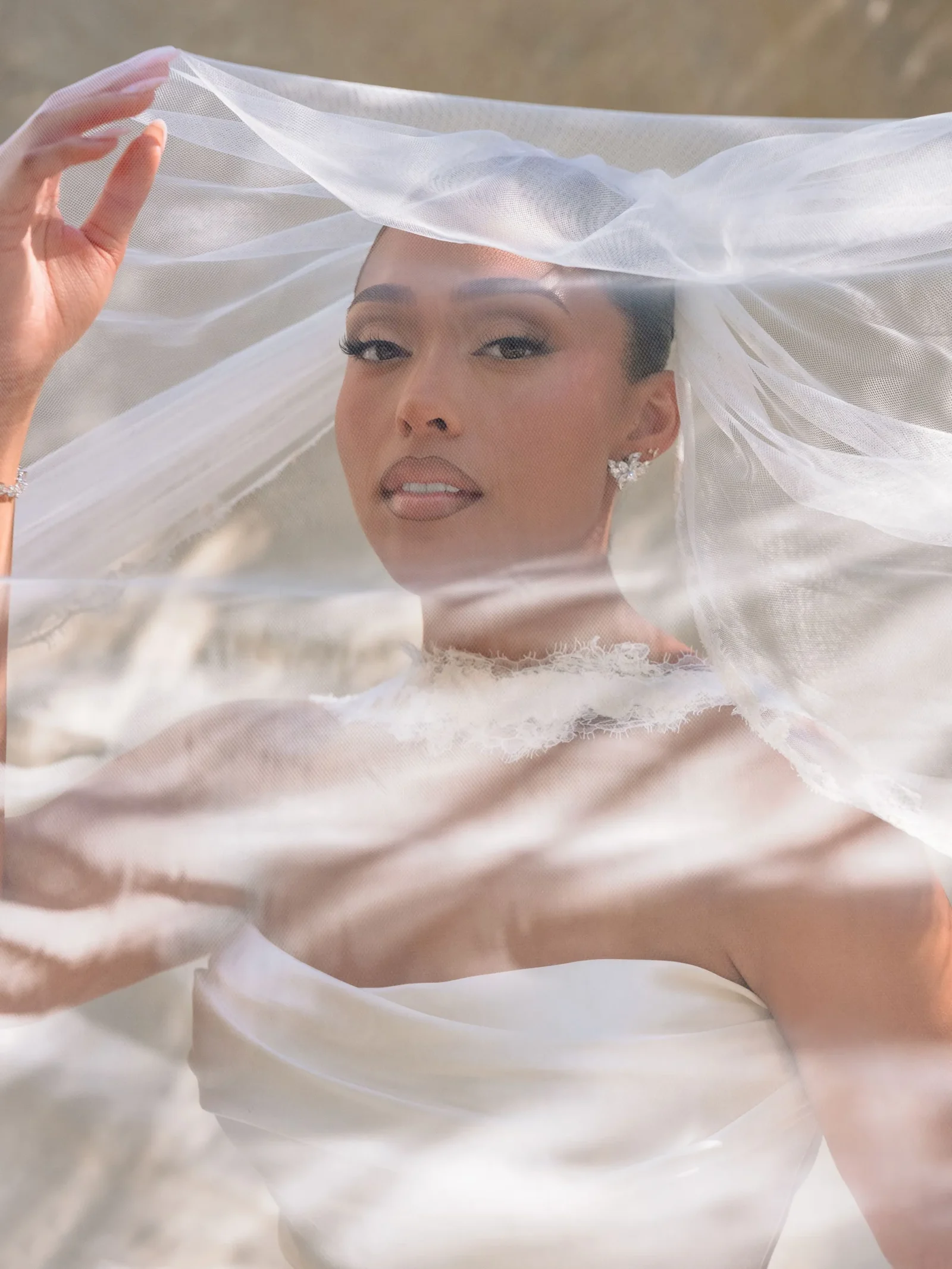 Close-up of a bride under a sheer veil, wearing a white lace wedding dress and diamond earrings, looking at the camera.