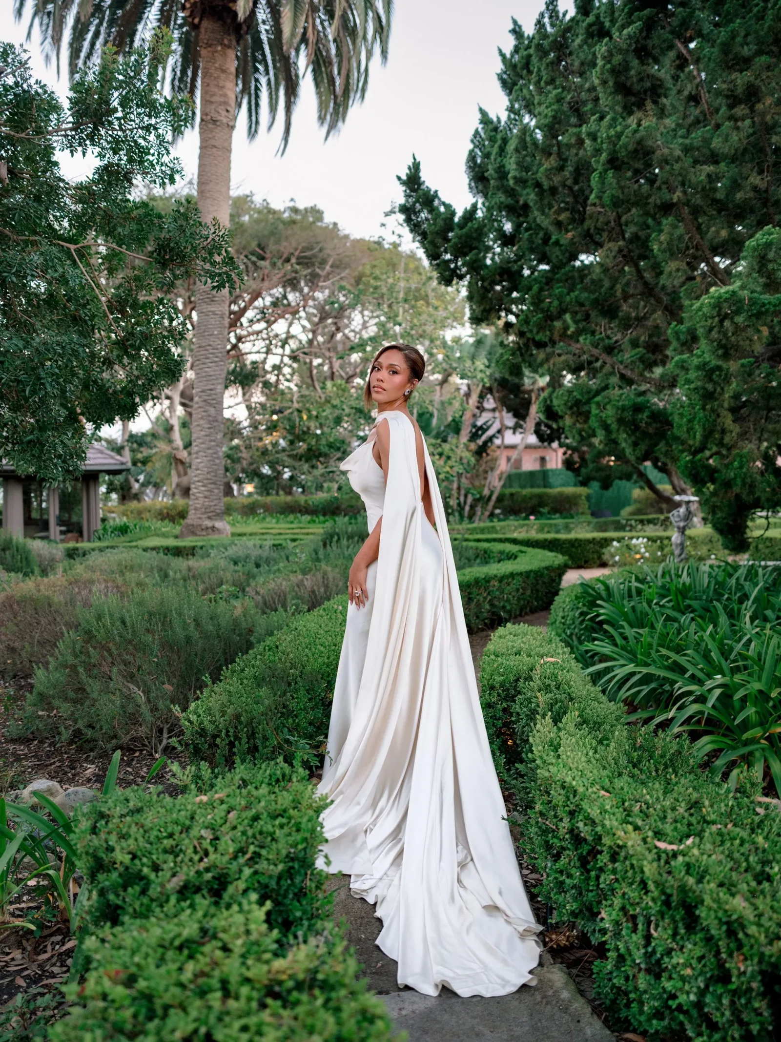 Woman in a long white gown posing on a stone path in a lush formal garden with trimmed hedges and trees.