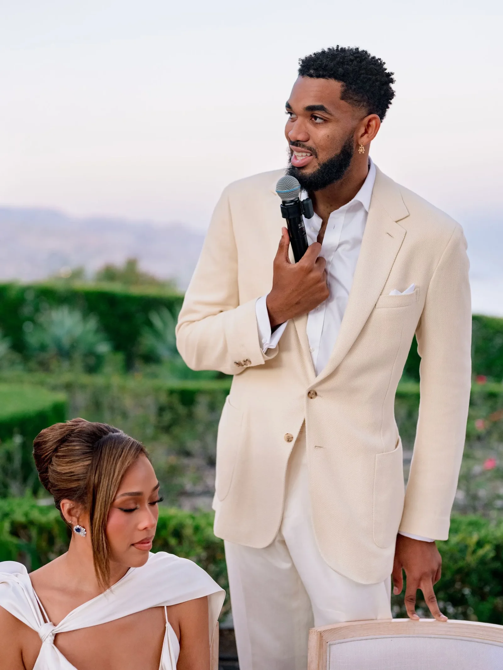 Man in a beige suit speaks into a handheld microphone outdoors, with a seated woman in the foreground.