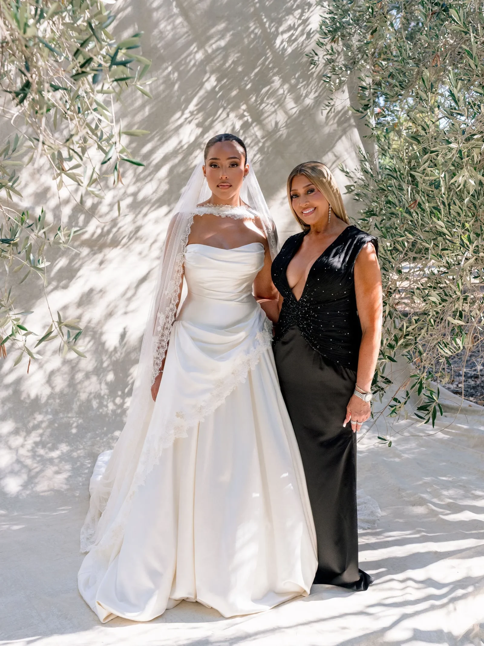 Bride in a white wedding gown with a lace veil standing next to a woman in a black evening gown outdoors among olive trees and dappled shade.