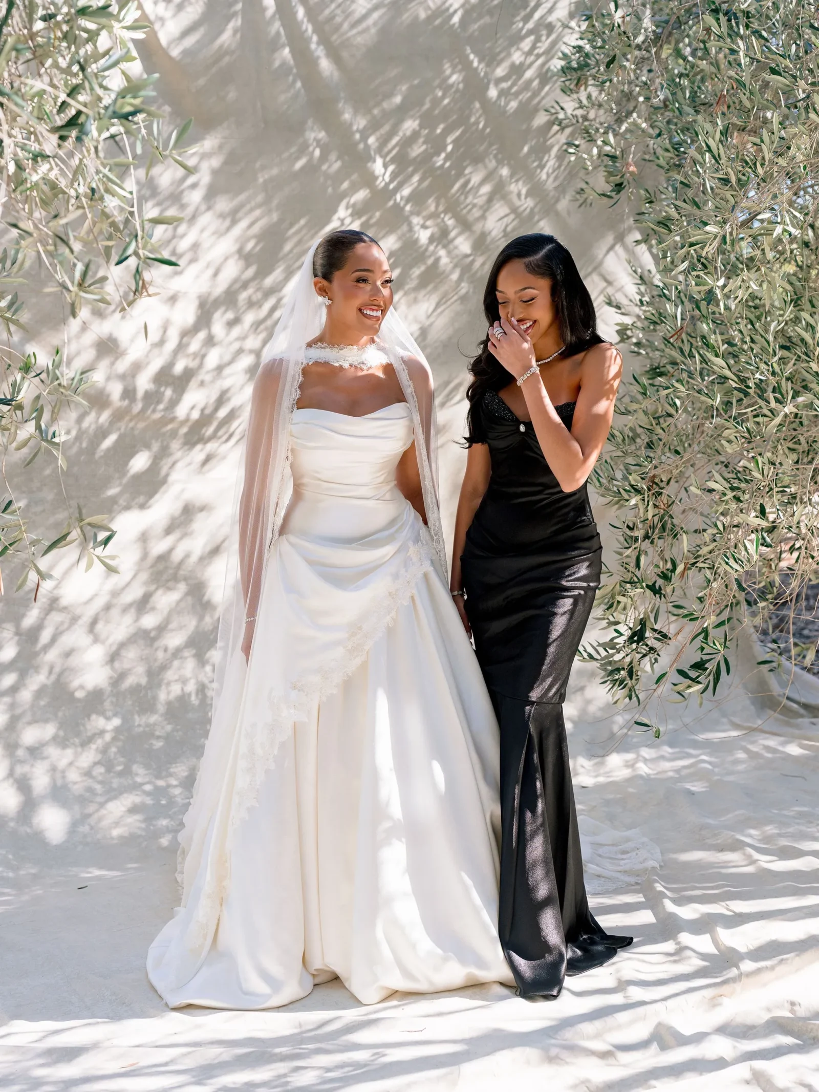 Bride in a white wedding gown with veil smiles next to a bridesmaid in a black dress, standing among sunlit olive trees.