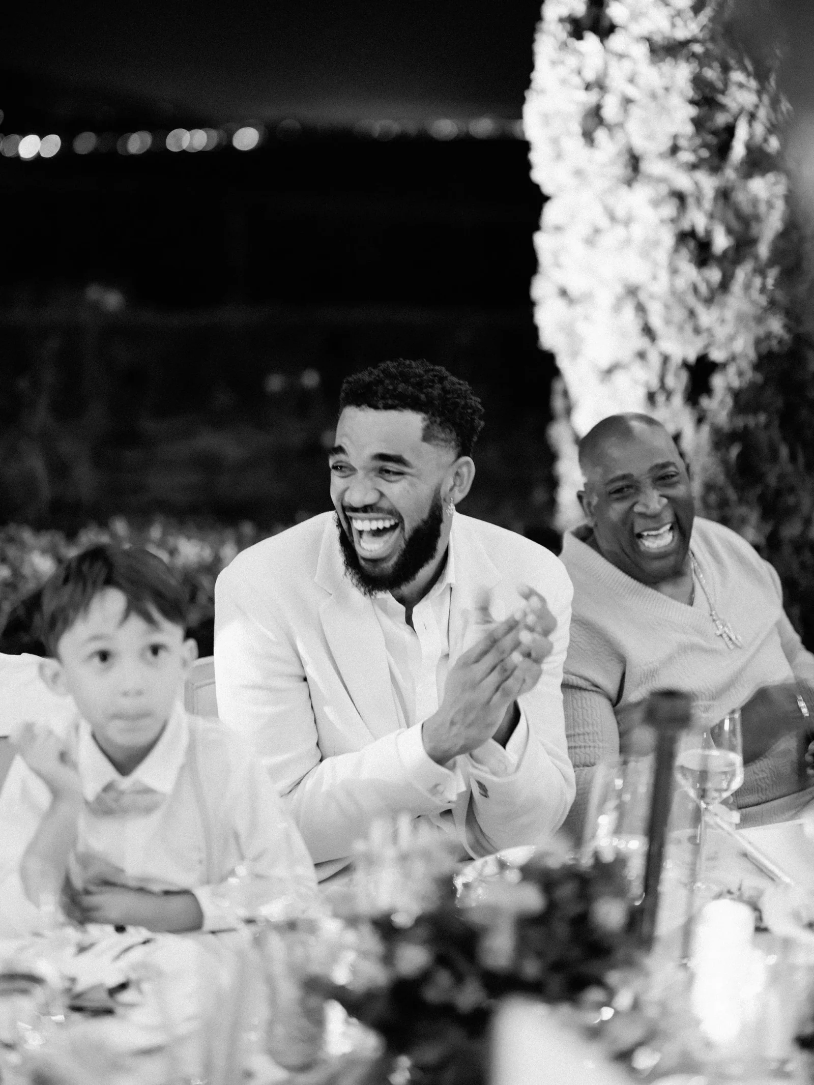 Bearded man in a light suit laughing at a dinner party, seated with a child and another smiling man beside floral decor.