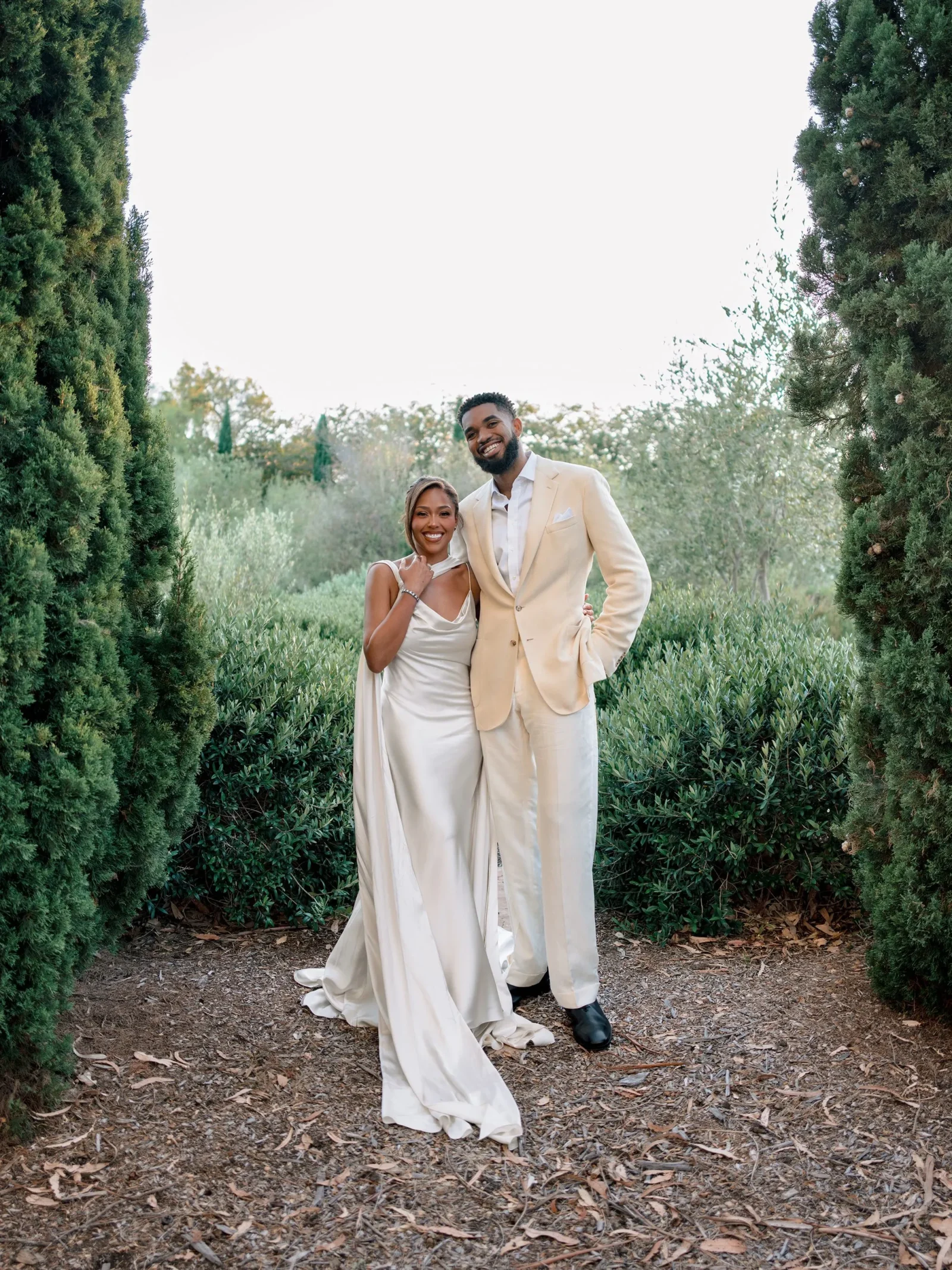 Smiling couple in cream dress and beige suit posing together in a sunlit garden framed by tall evergreen hedges.