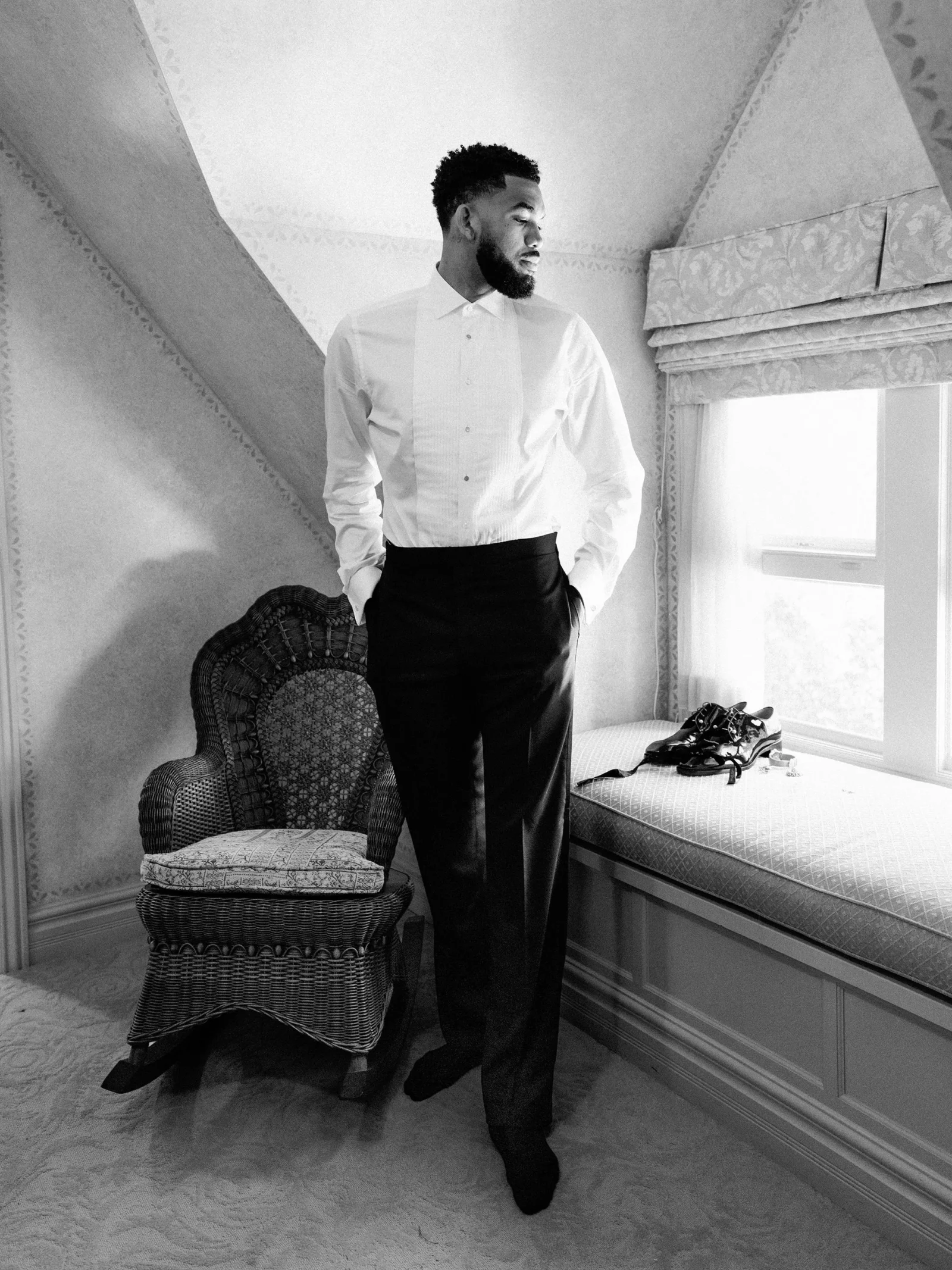 A man in a white dress shirt and dark trousers stands in a vintage, carpeted room beside a wicker chair and a window seat with shoes on it.