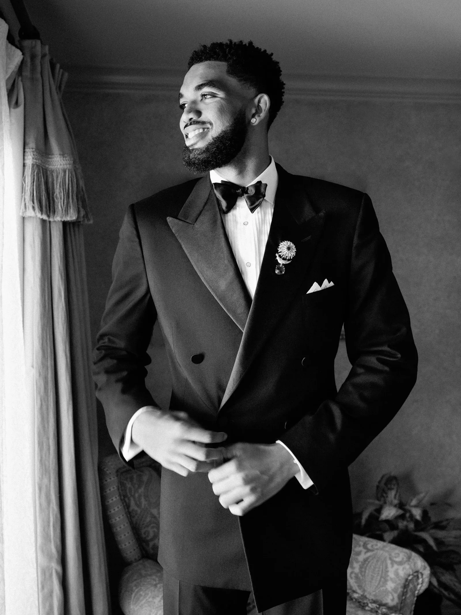 Black-and-white portrait of a man in a tuxedo adjusting his cufflinks beside a window, wearing a bow tie and boutonniere and smiling.