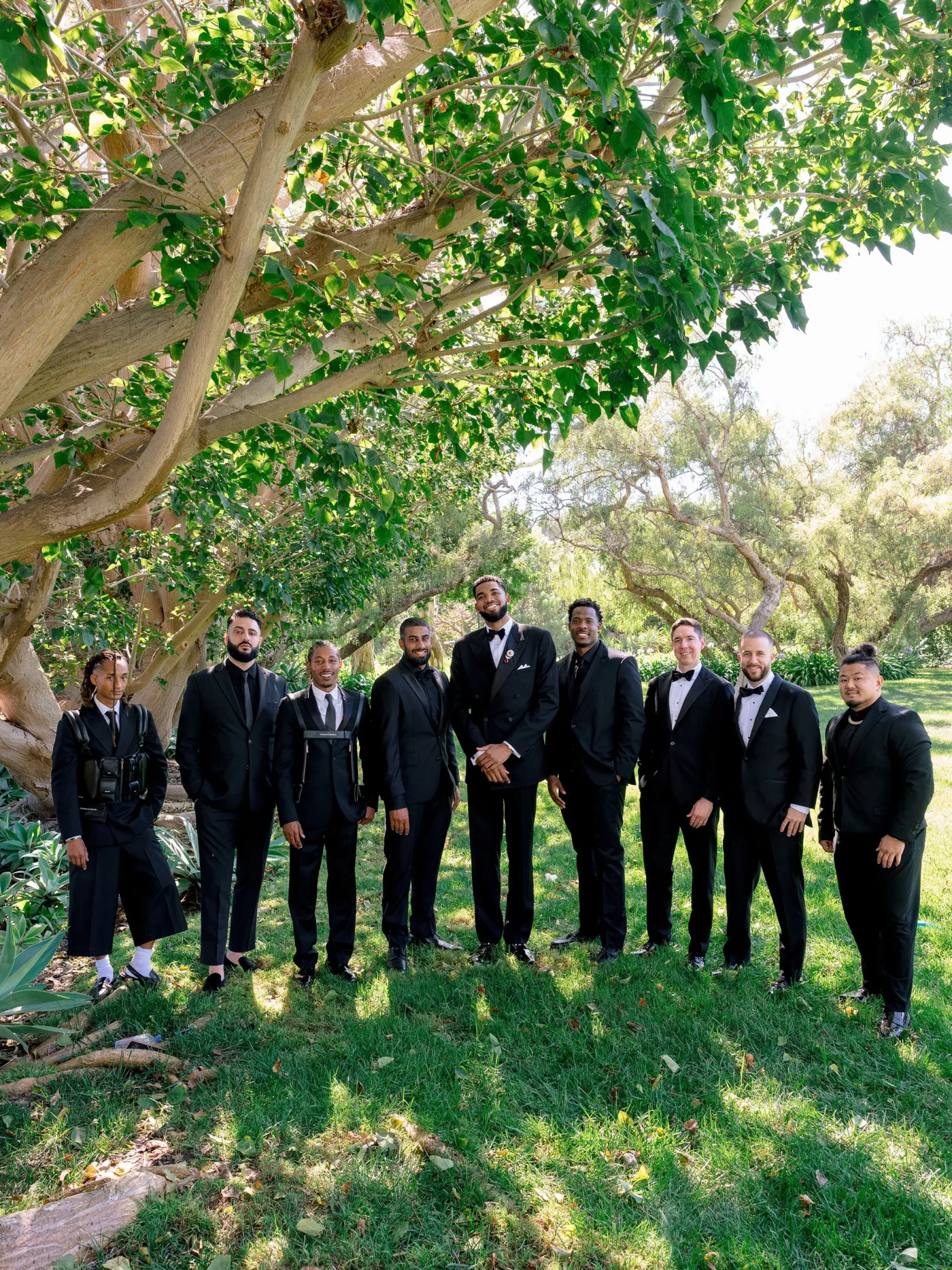 Group of men in black suits standing in a park under a large tree in bright daylight.