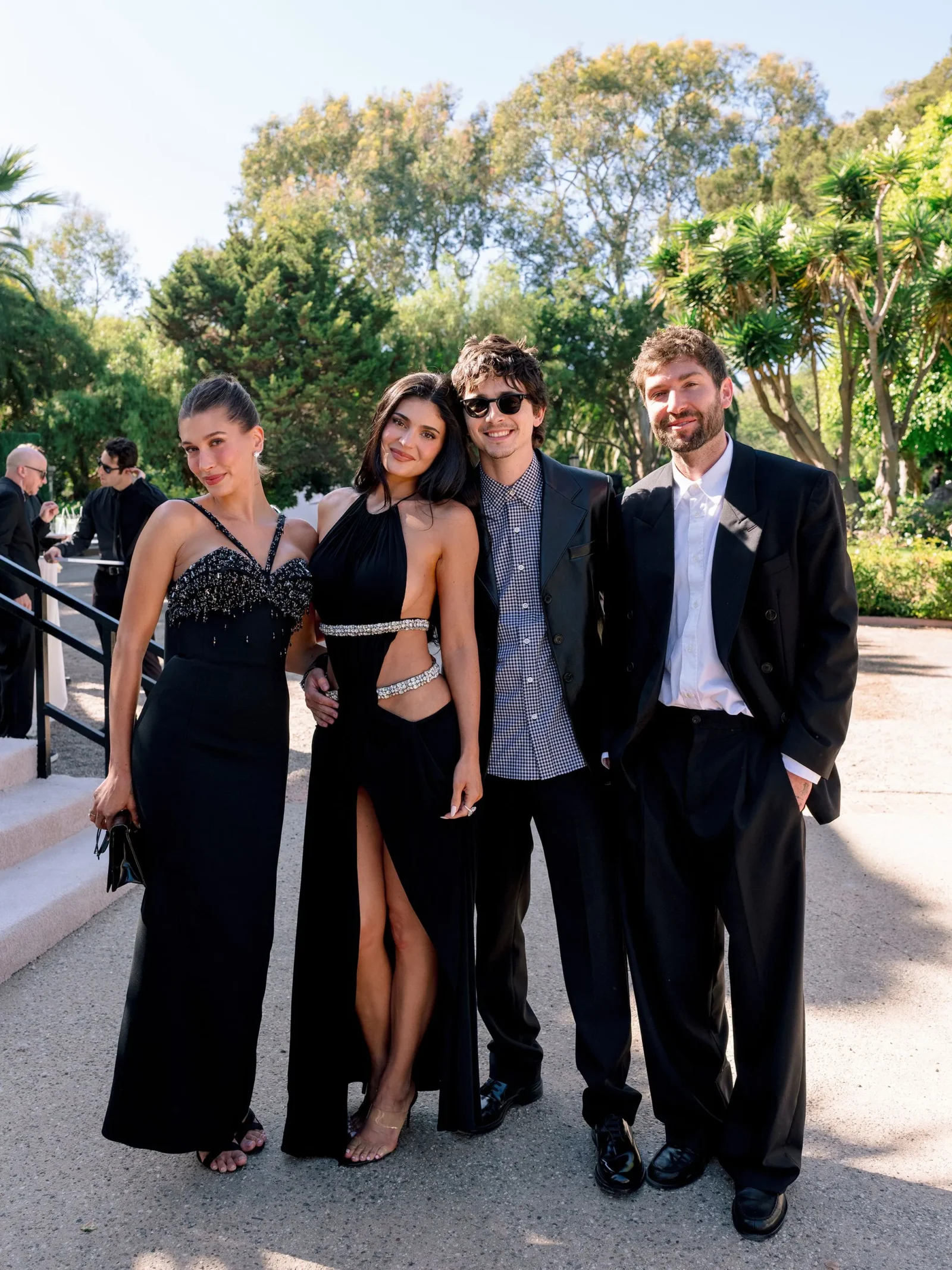 Four friends dressed in formal black attire pose together outdoors on a sunny day, smiling at the camera.