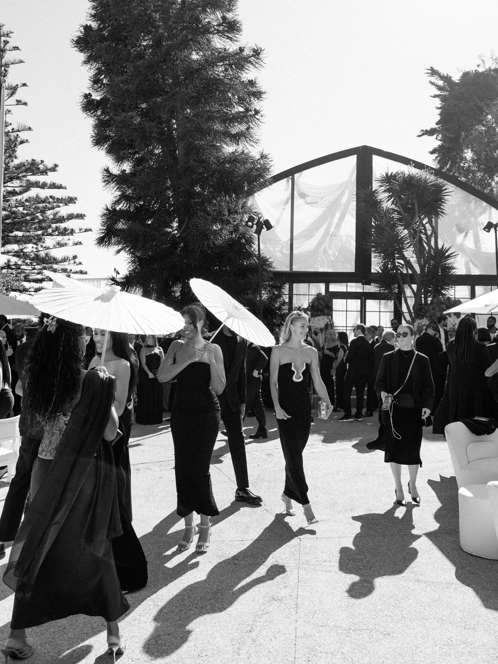 Outdoor formal event with guests in black attire under white umbrellas, casting long shadows near a glass greenhouse structure.