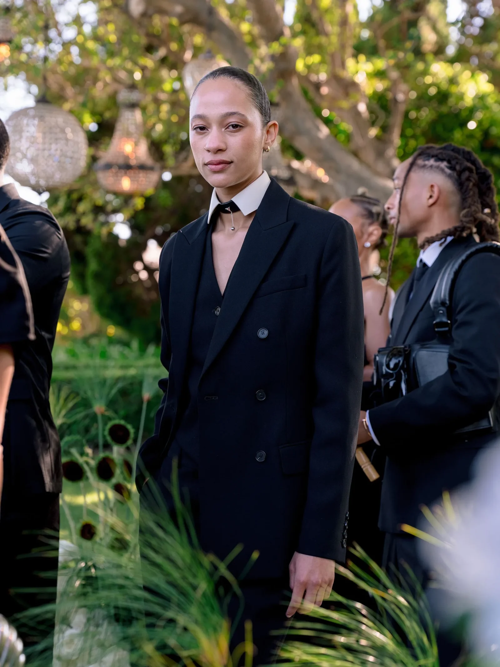 Person in a black double-breasted suit standing outdoors among greenery, facing the camera with a calm expression.