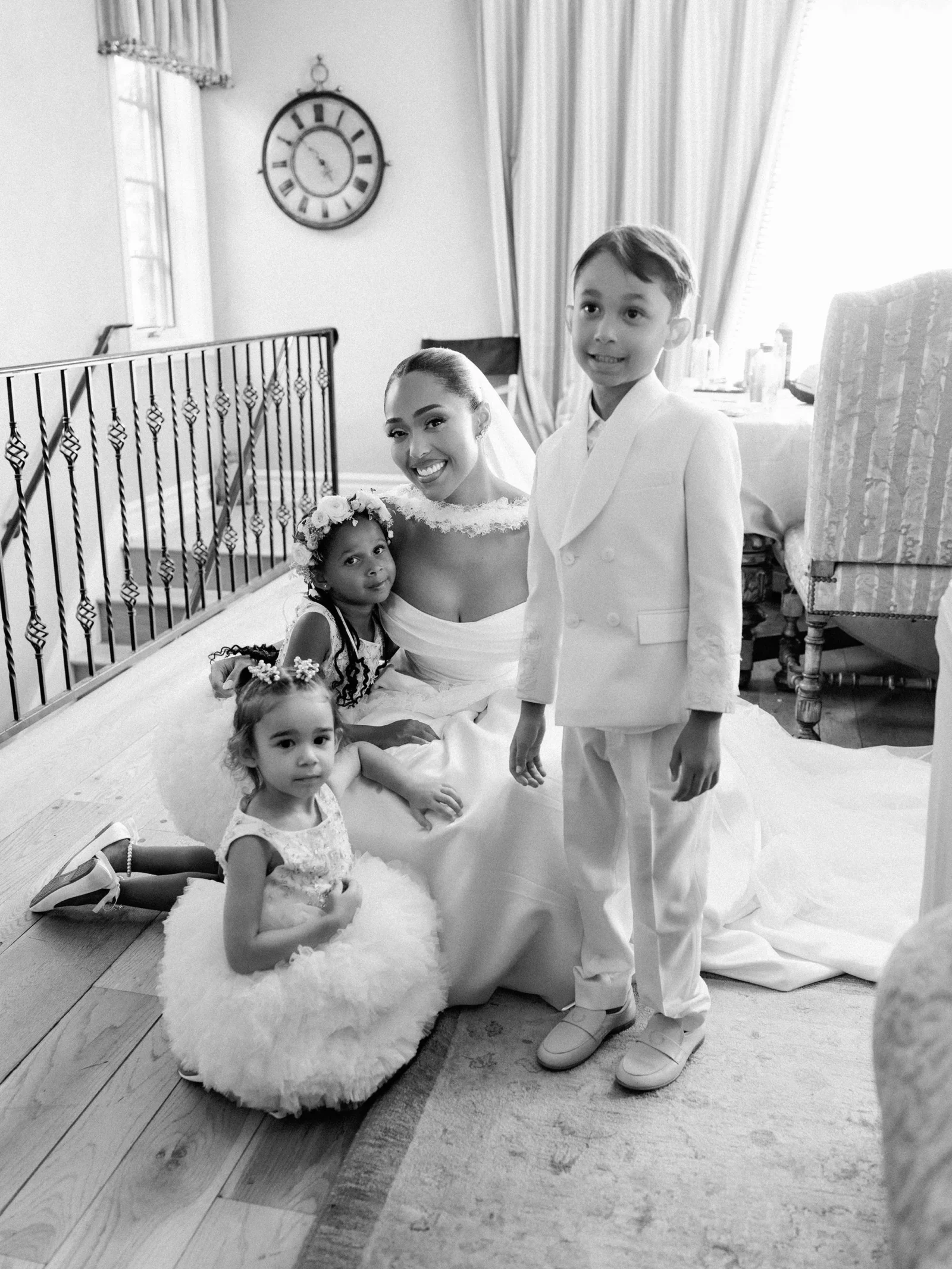 Bride in a wedding dress kneels with three young girls and a boy in formal attire indoors, clock on the wall in the background.