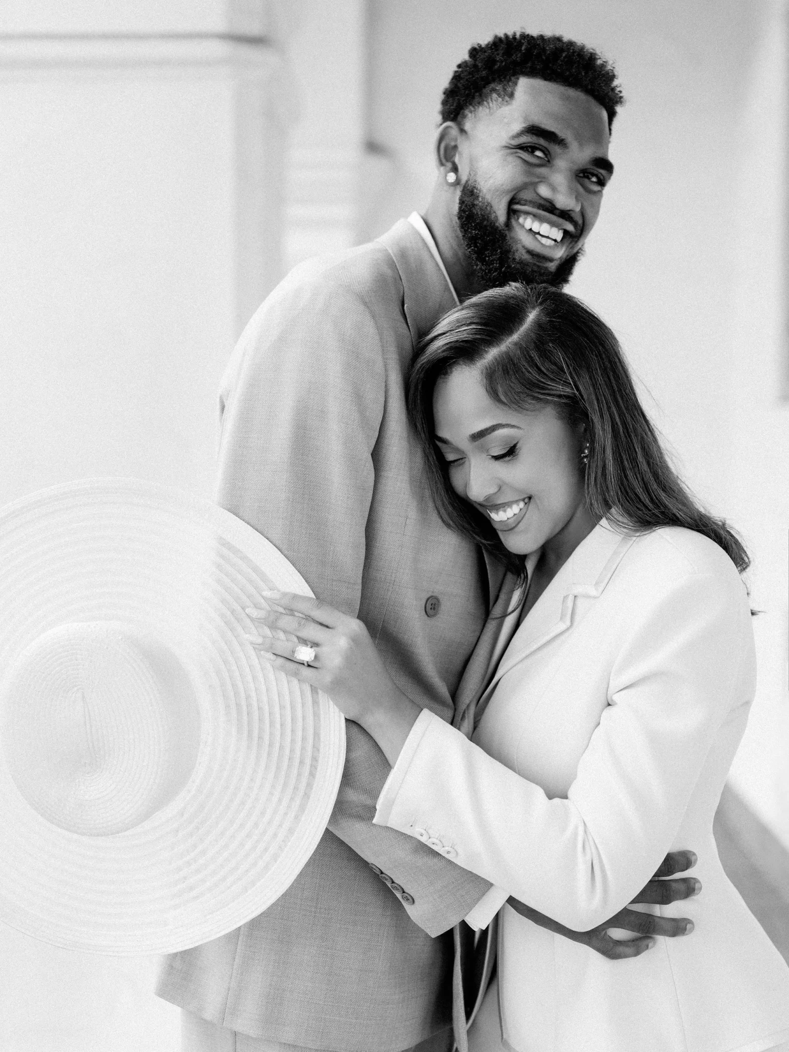Smiling couple hugging; man in a blazer with a beard, woman resting her head on his chest, a wide-brimmed hat visible in the foreground.