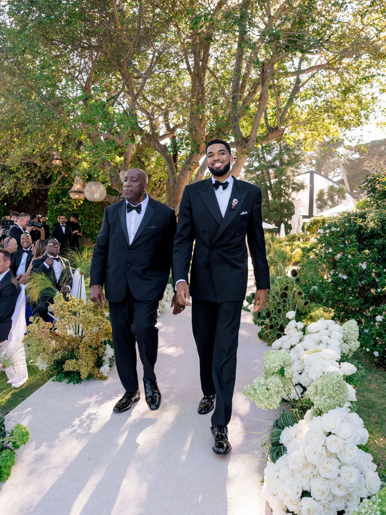 Two men in black tuxedos walk down an outdoor wedding aisle lined with white roses and greenery, smiling as guests watch in the background.