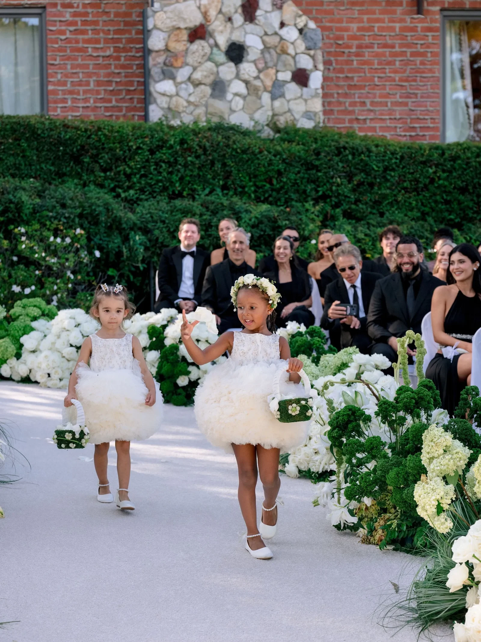 Two young flower girls in white fluffy dresses walking down a floral aisle with baskets, during a formal outdoor wedding.
