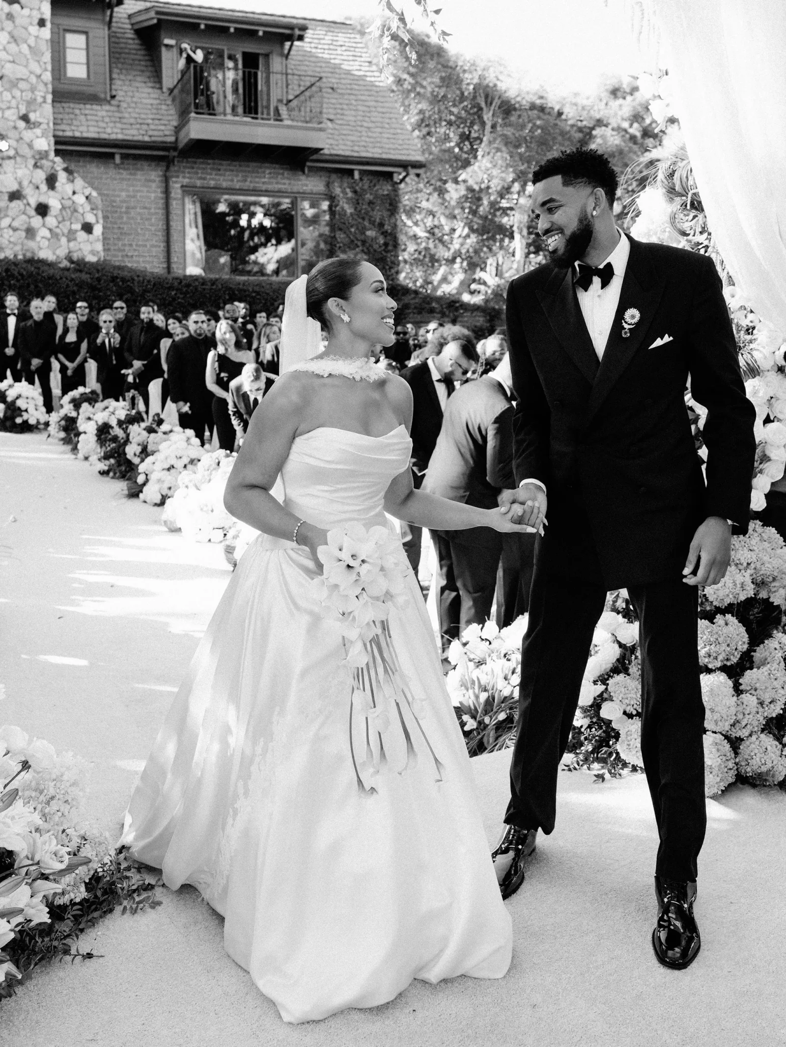 Bride in a strapless gown with veil holds a bouquet as she walks hand in hand with a smiling groom in a black tuxedo at an outdoor wedding, with guests and floral decorations in the background.