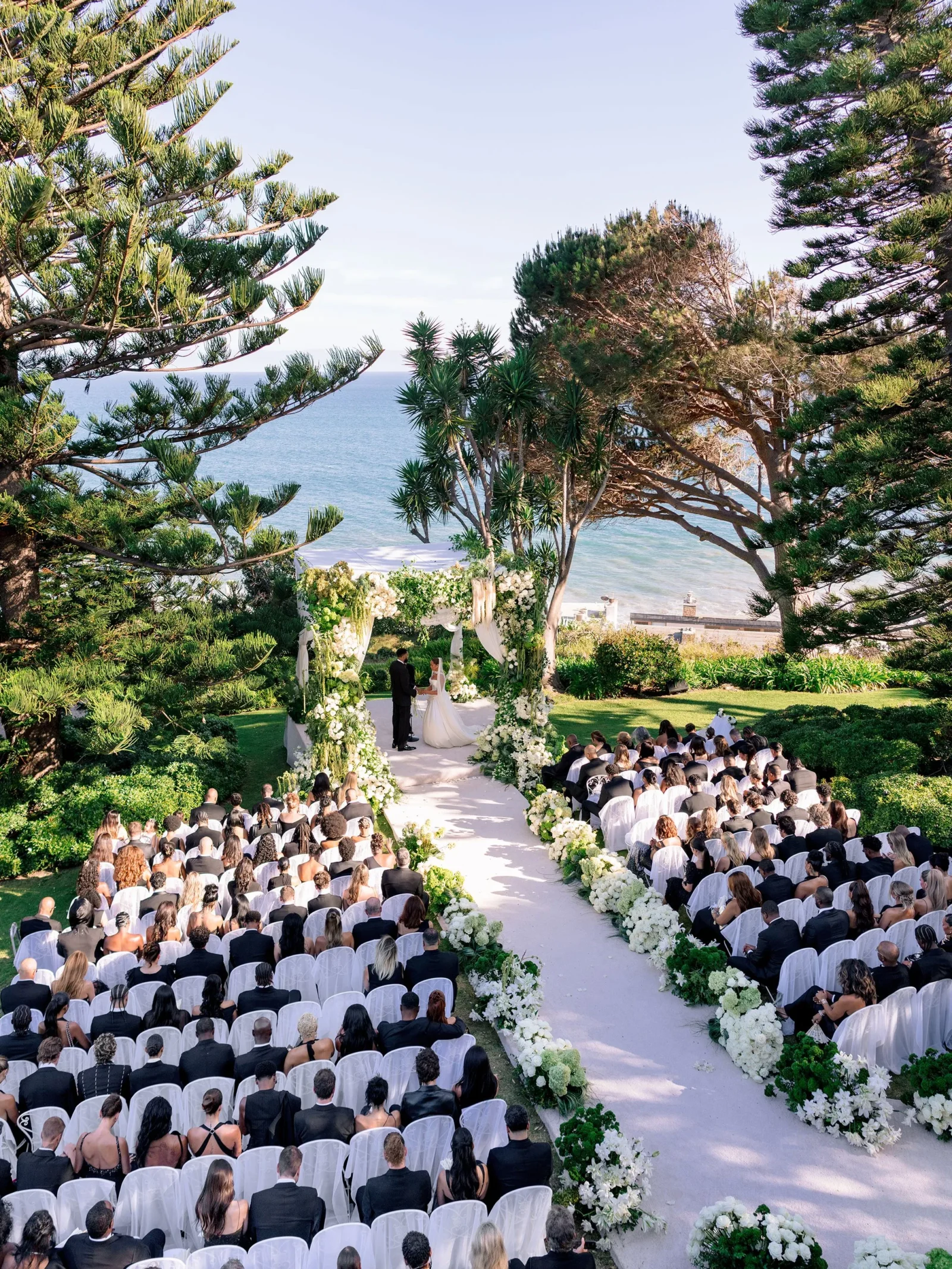 Bride and groom exchange vows under a white floral arch on a lawn by the sea, with guests seated and watching.