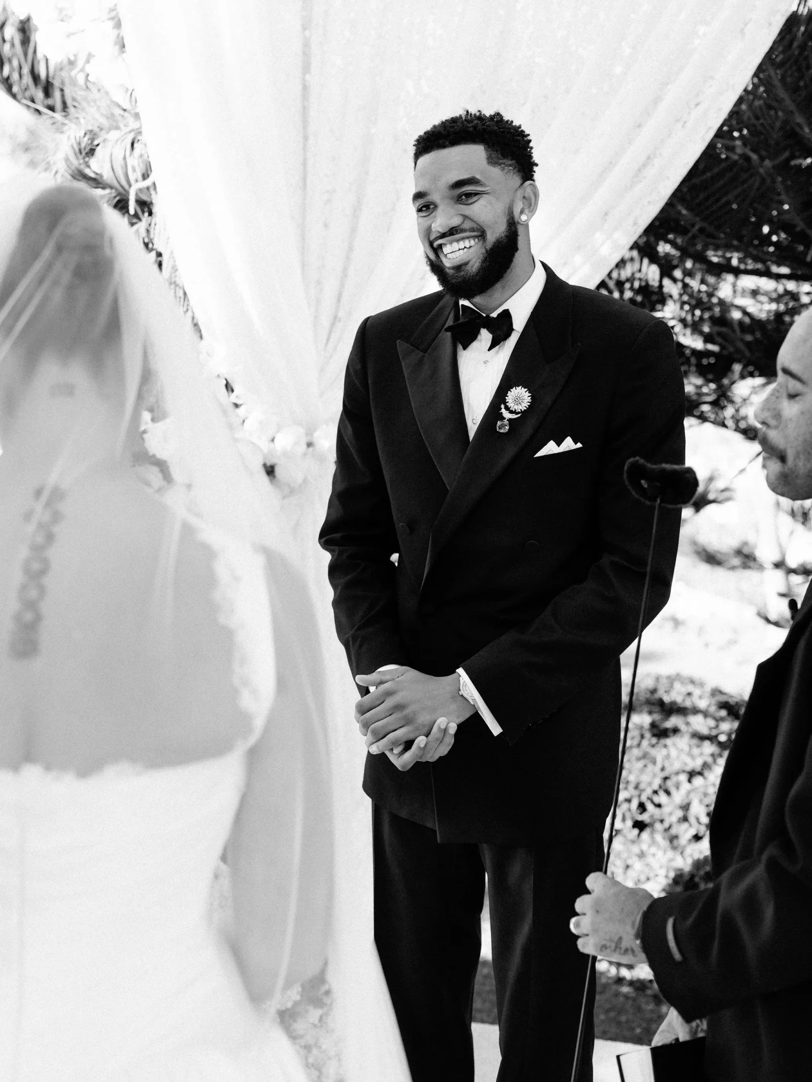 Smiling groom in a black tuxedo with bow tie and boutonniere, standing beside a bride in a wedding dress.