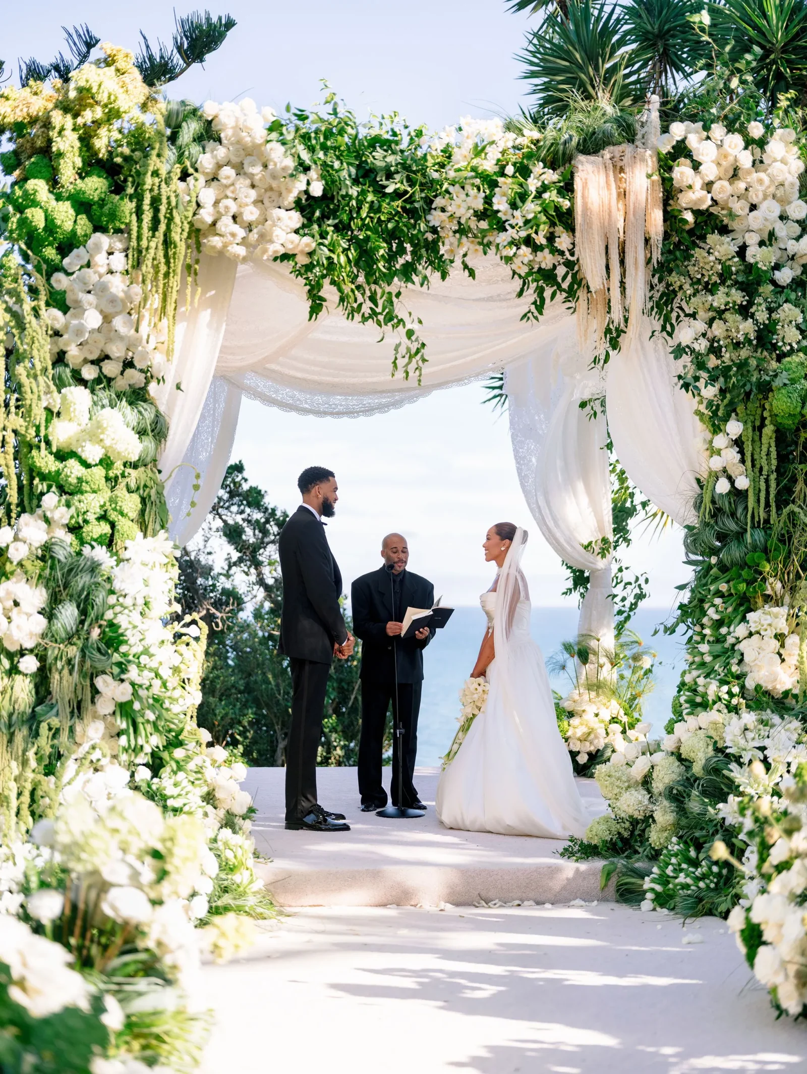 Bride and groom stand under an ornate white floral arch with greenery, exchanging vows as officiant reads nearby by the seaside.