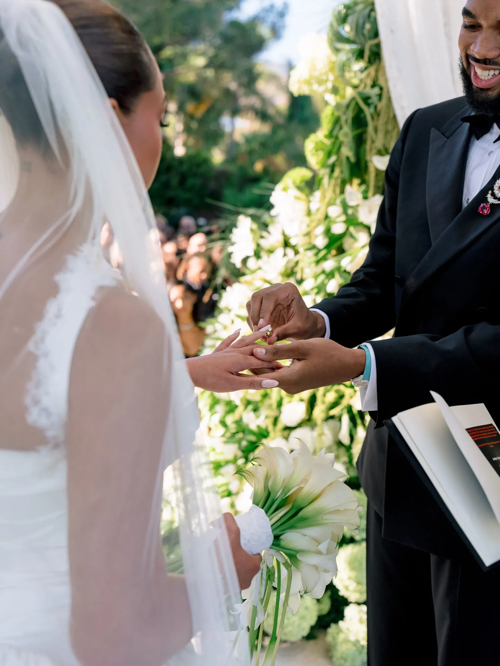 Bride and groom exchange rings during an outdoor wedding, with the bride holding a white bouquet and veil visible on the left.