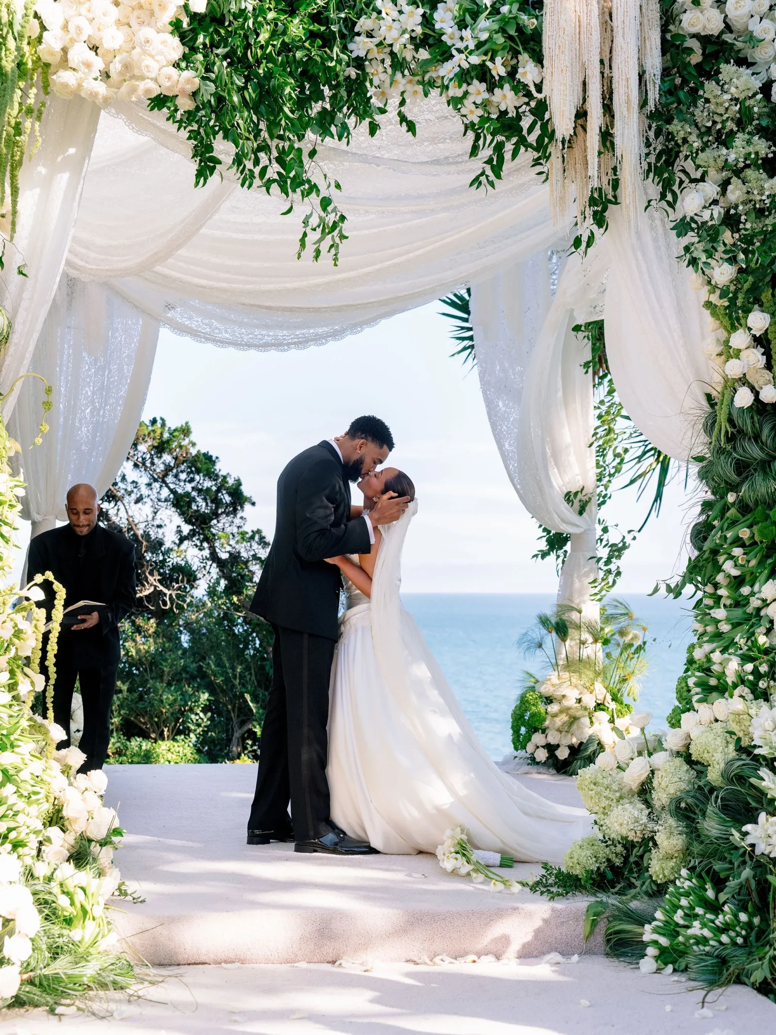Bride and groom kissing under a white floral arch by the seaside, with greenery and ocean in the background.