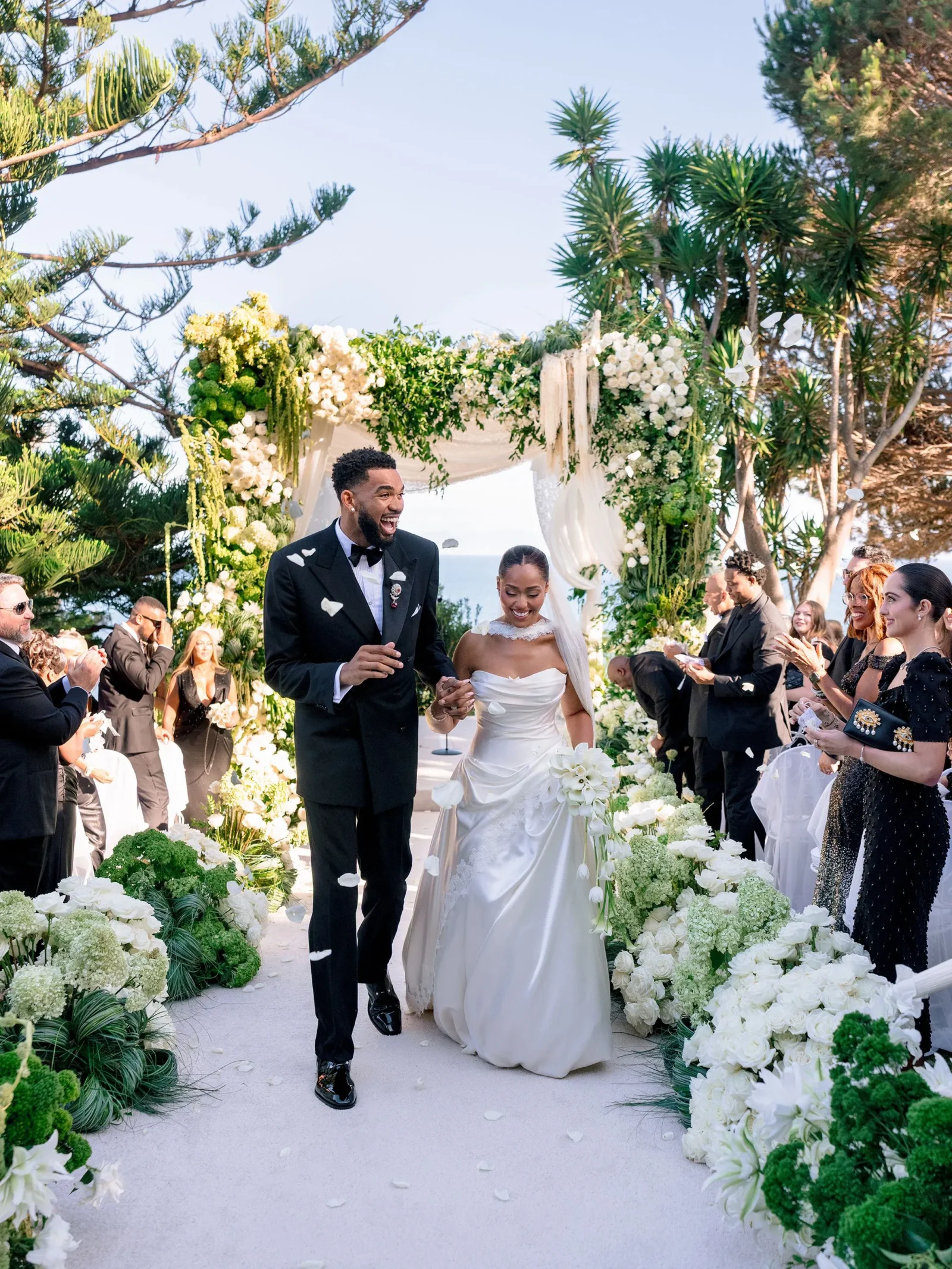 Bride and groom walking down a flower-adorned outdoor wedding aisle amid guests, beneath a white floral arch near the sea.