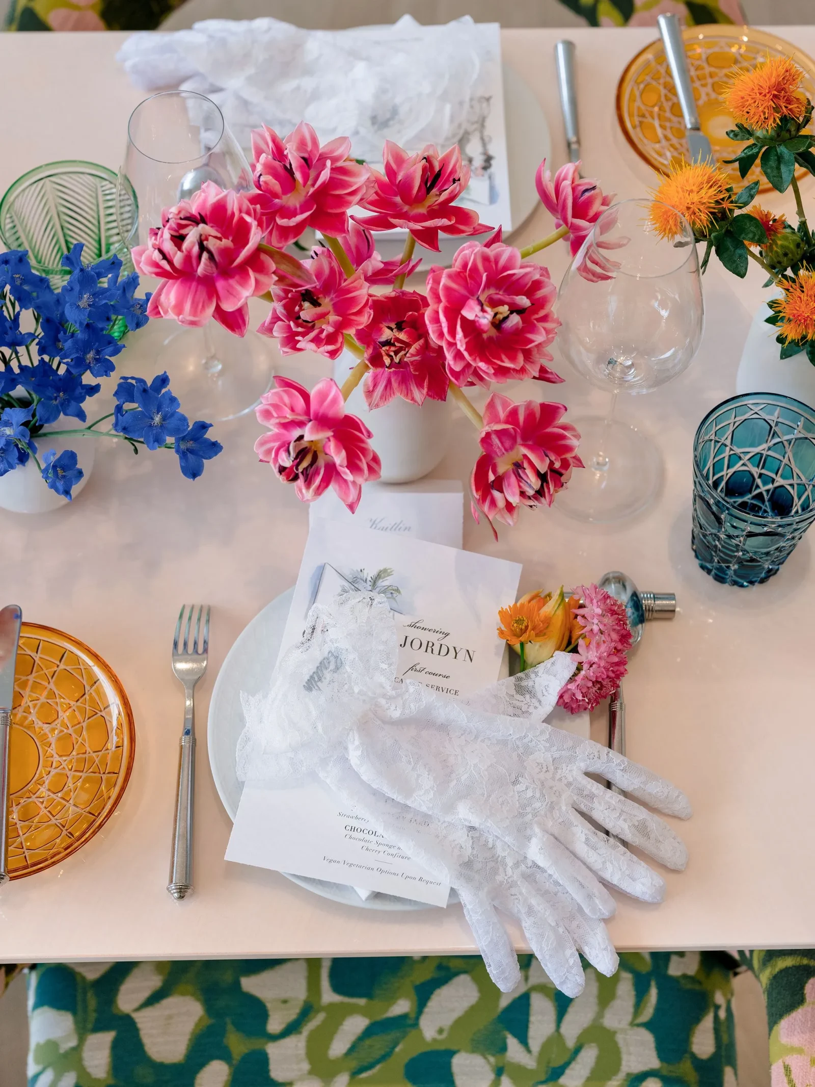Table setting with pink and blue flowers, white lace gloves, and orange patterned plates with wine glasses in a bright dining setup.