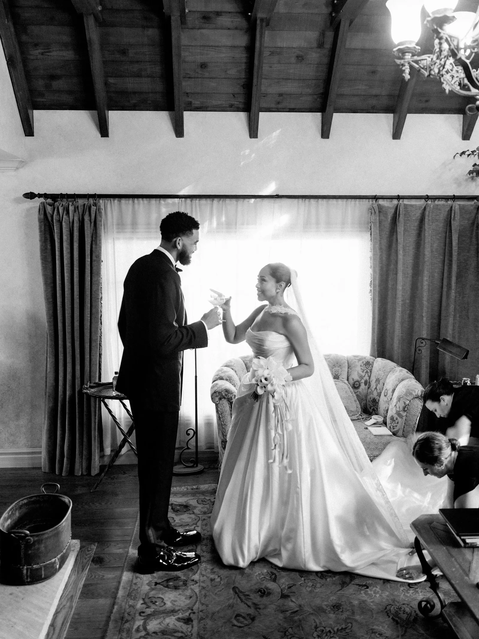 Bride in a strapless wedding dress toasts groom in a dark suit in a sunlit, rustic living room with a floral sofa in the background.