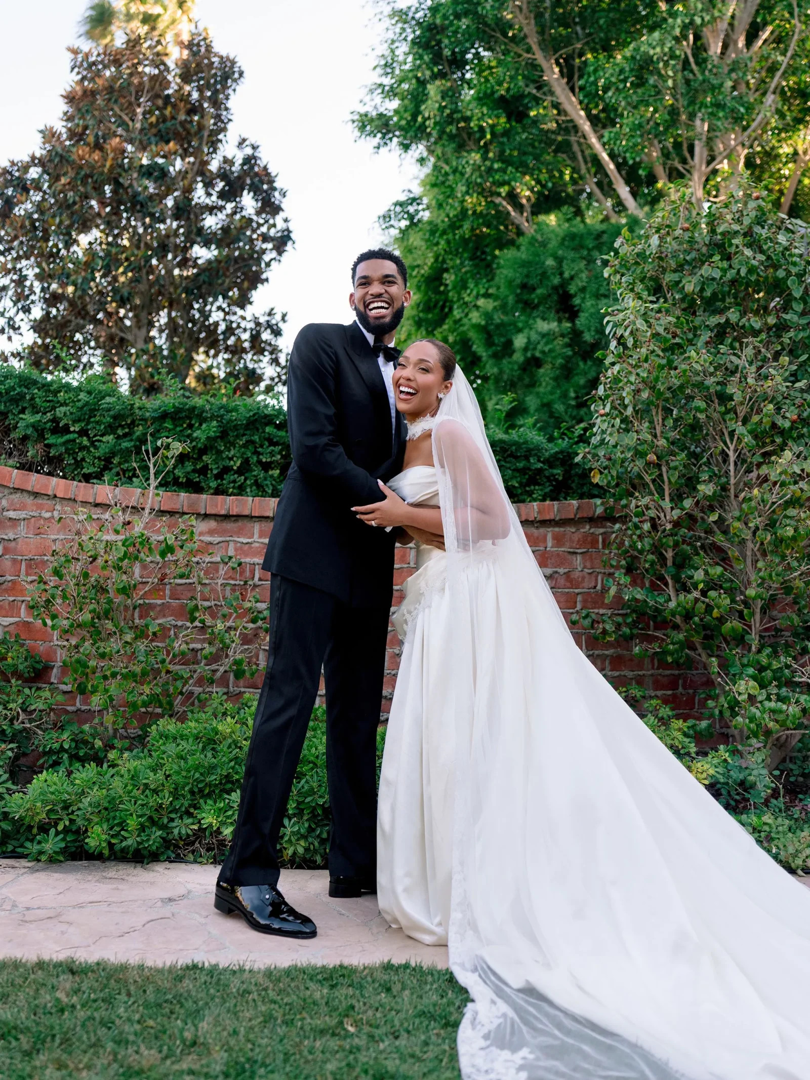 Bride in a white wedding gown with a veil smiling as she hugs her groom in a black tuxedo outdoors, brick wall and greenery behind them.