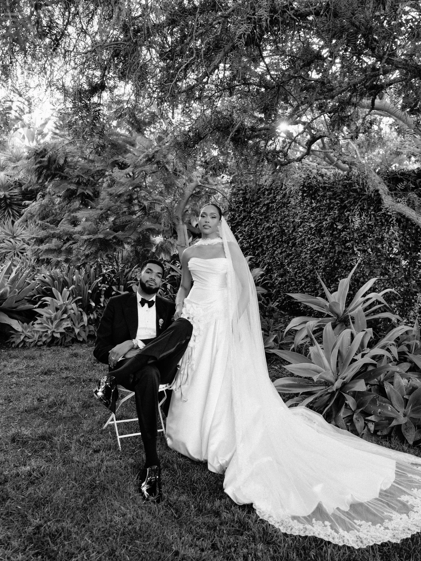 Bride in a strapless wedding gown with veil stands beside a seated groom in a suit in a lush garden setting.