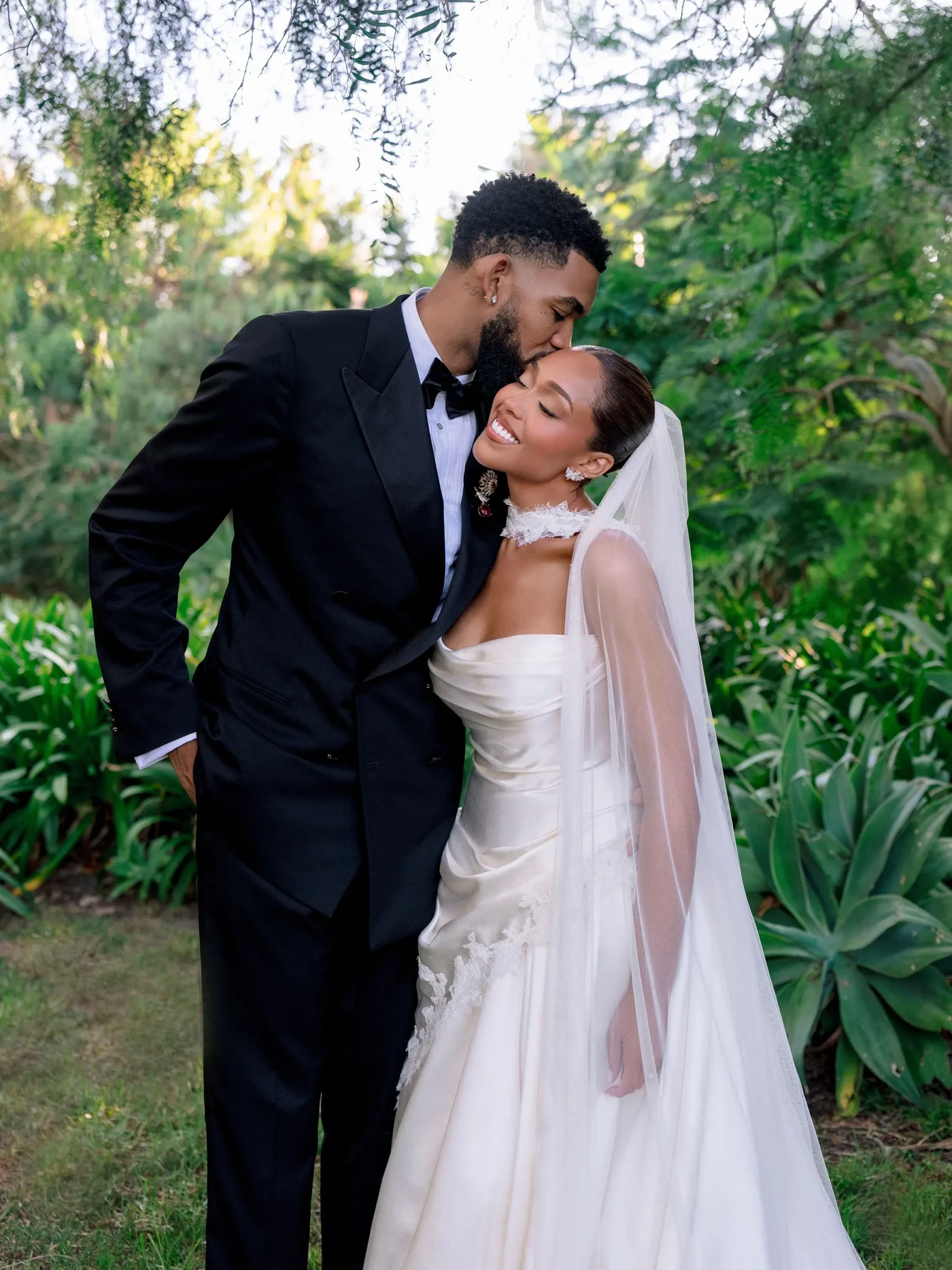 Bride in a white wedding gown and veil smiles as a groom in a black tuxedo kisses her forehead in a garden setting.
