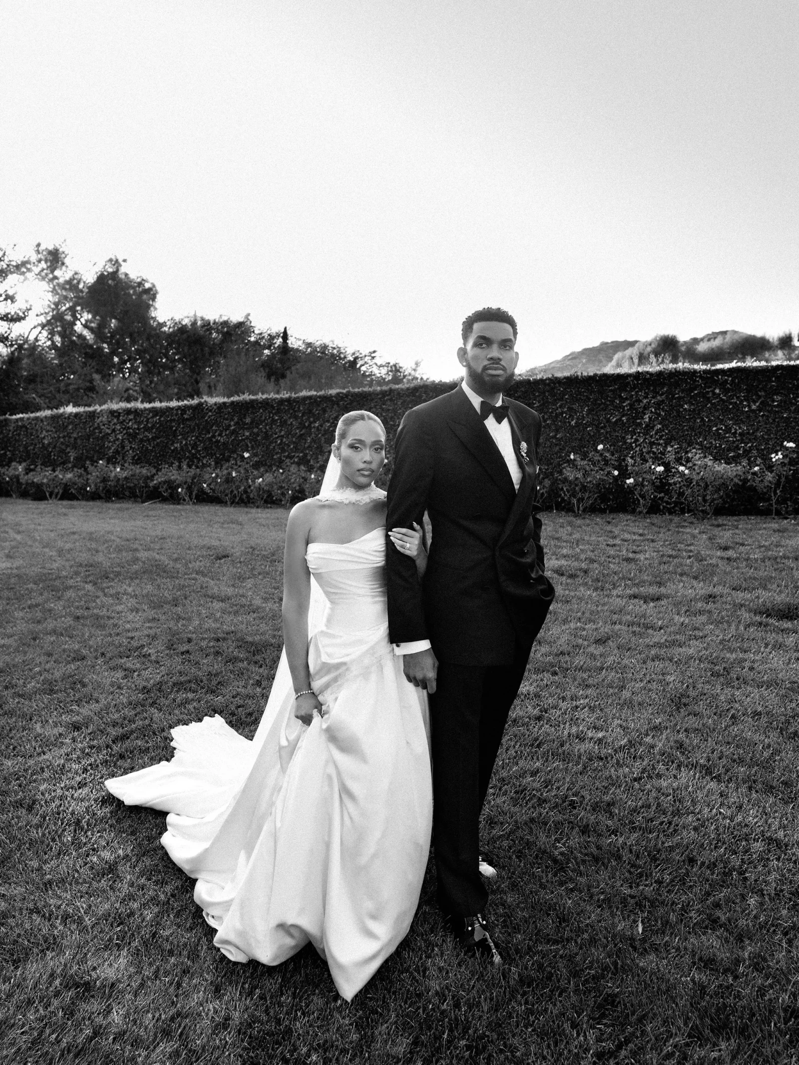Bride and groom in wedding attire stand arm in arm on a grassy lawn with a hedge in the background; black-and-white photo.
