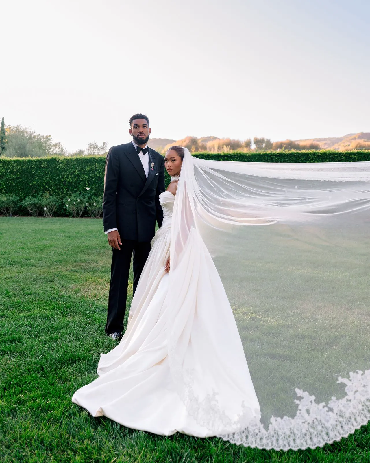 Bride and groom pose on a green lawn; bride’s flowing veil extends behind them.