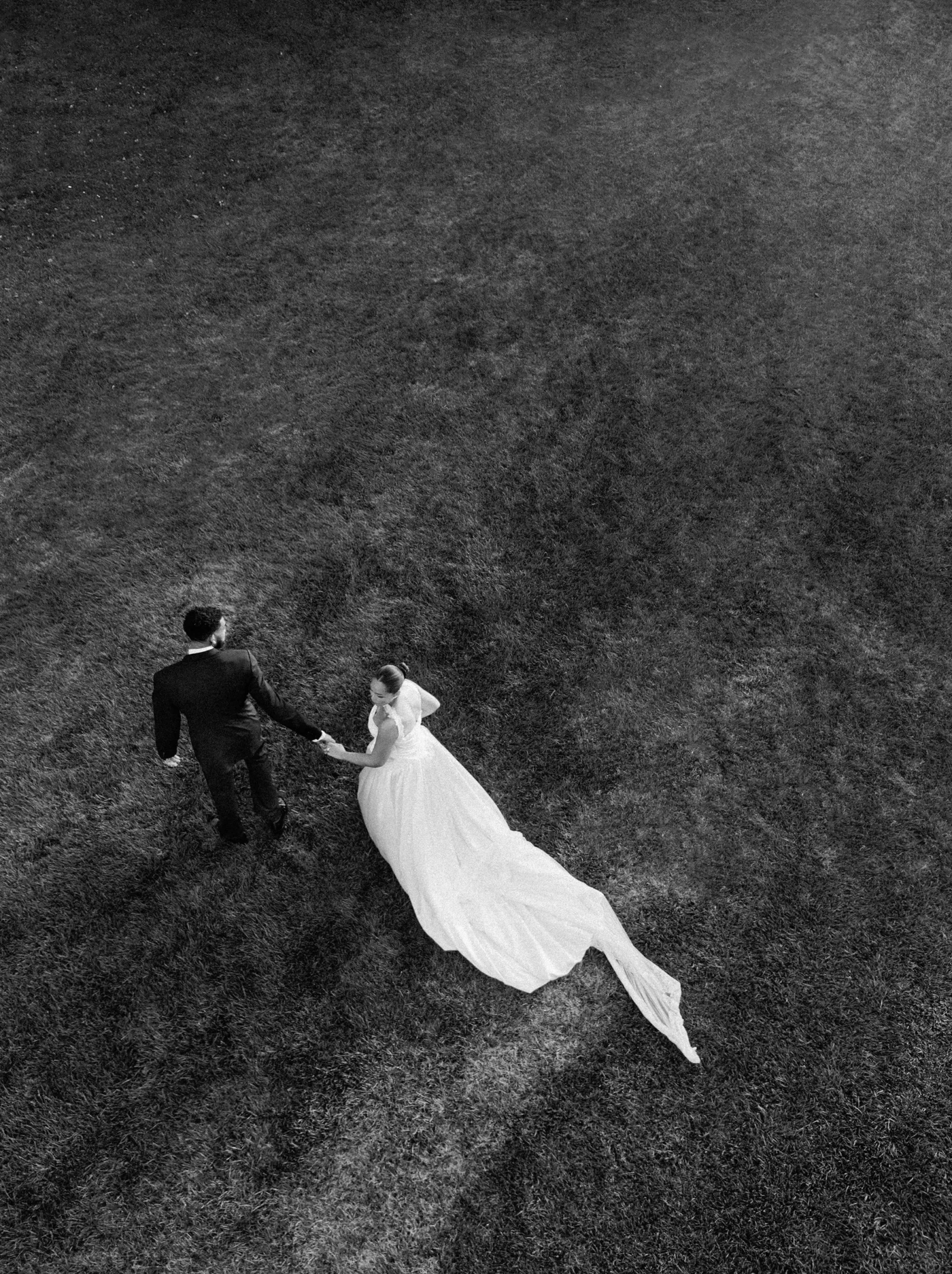 Aerial view of a bride in a white wedding dress and a groom in a dark suit walking hand in hand across a grassy field.